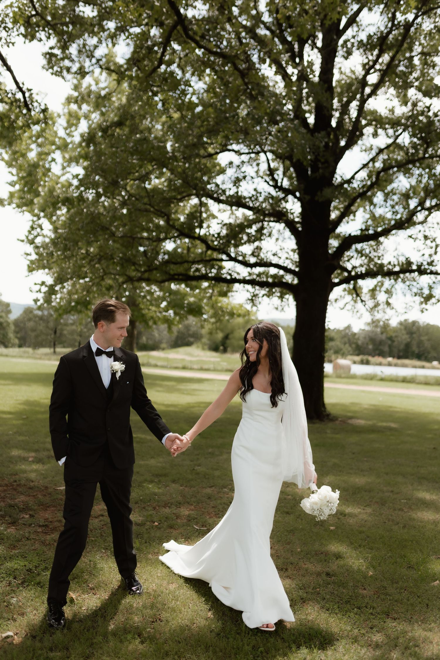 Bride and groom hold hands while walking across a green lawn with a large oak tree behind them.