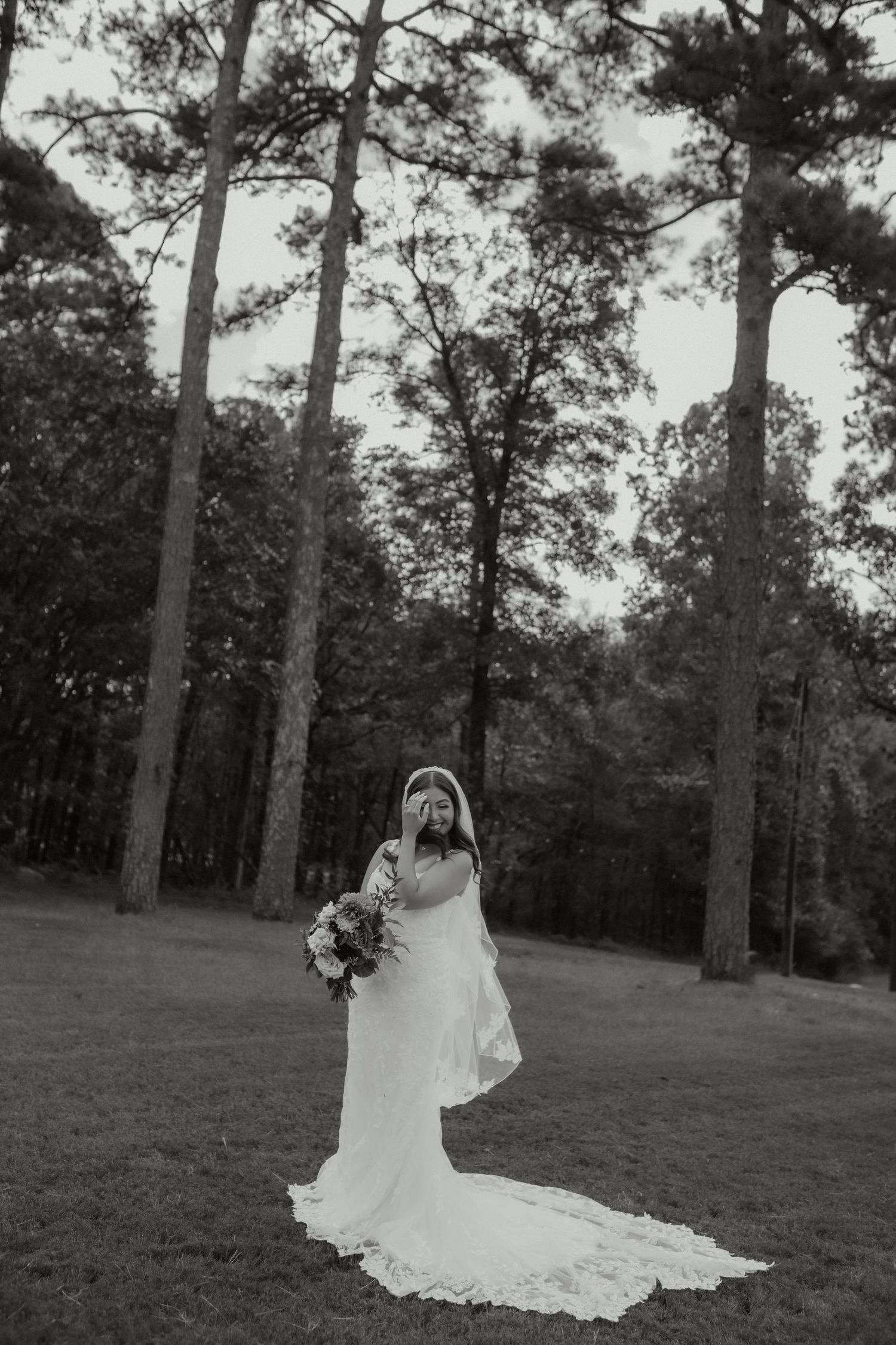 Black and white portrait of a bride in a flowing gown and veil standing among tall pine trees.