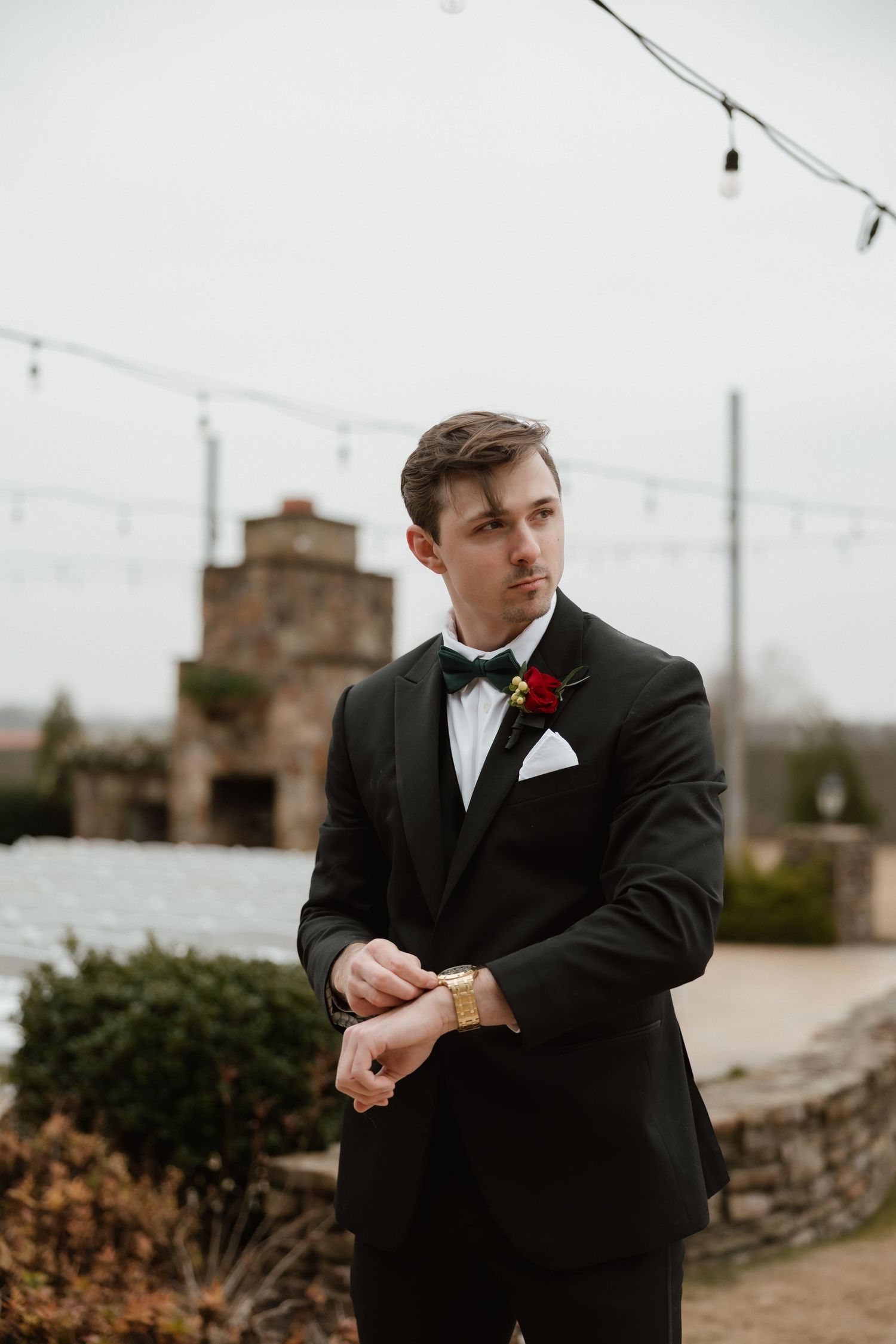 Groom in black tuxedo with red boutonniere adjusting cufflinks outdoors at wedding venue.