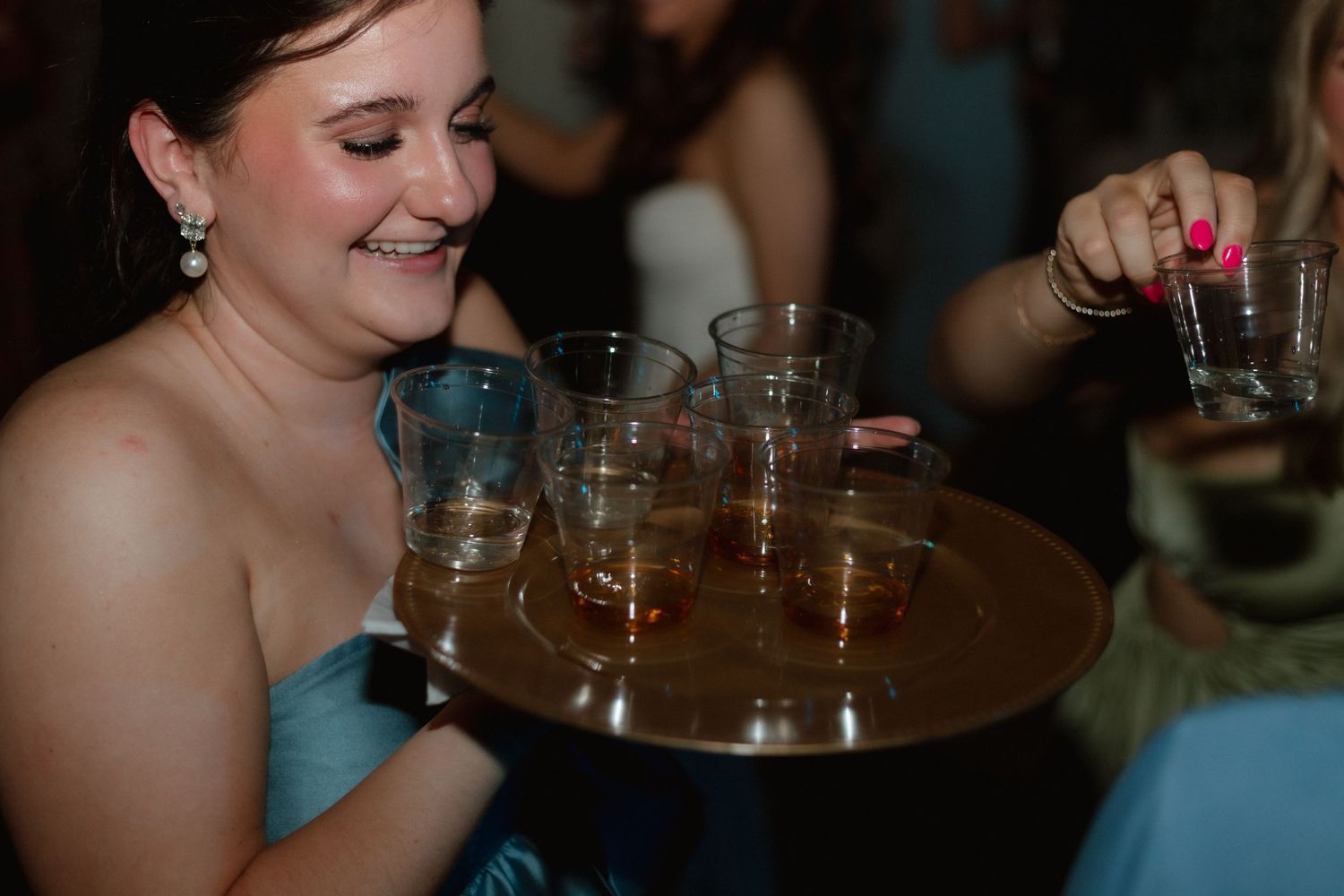 Bridesmaid in teal dress carrying a tray of whiskey shots at a wedding reception.
