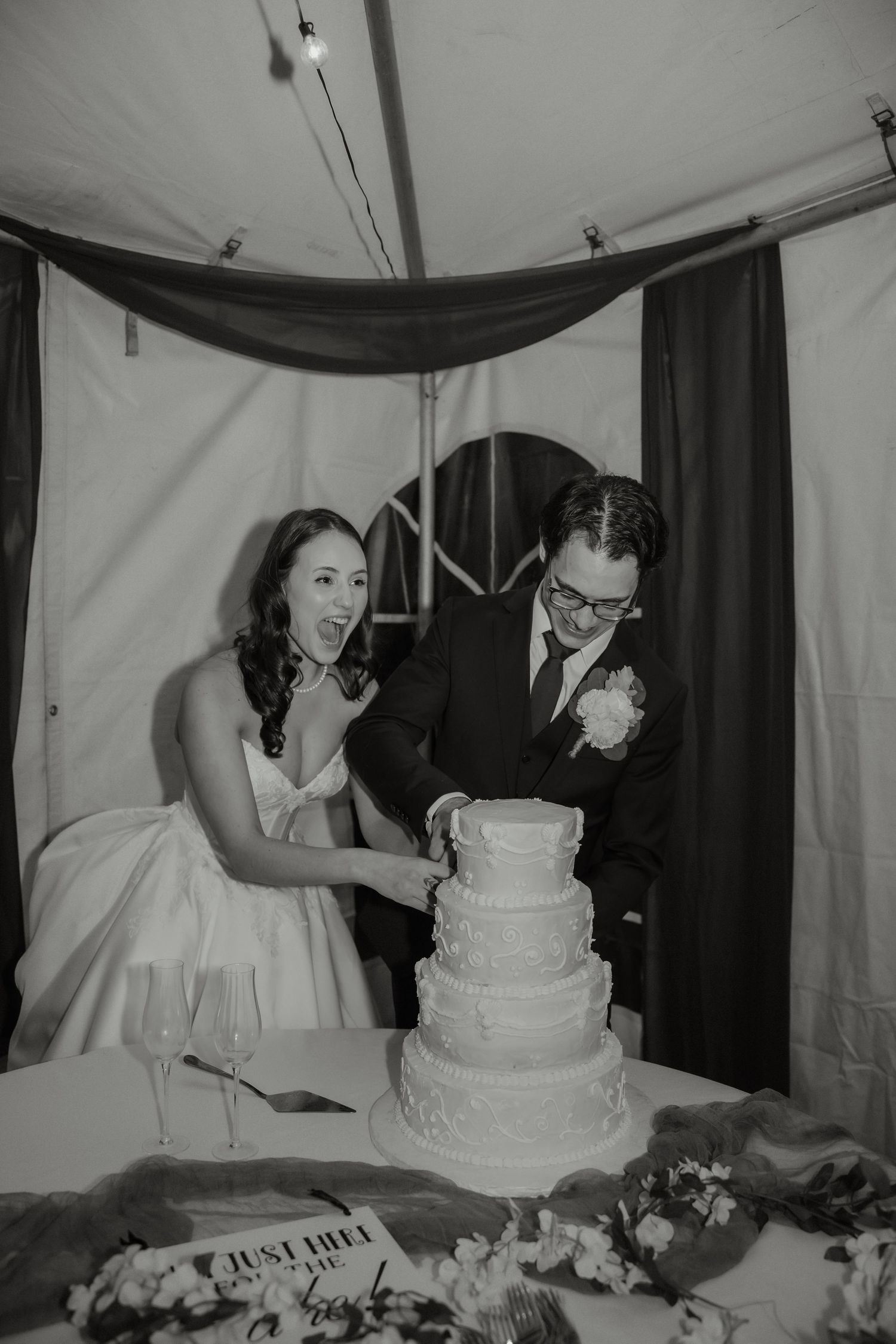 Excited bride and groom cutting tiered white wedding cake at reception in black and white photo.