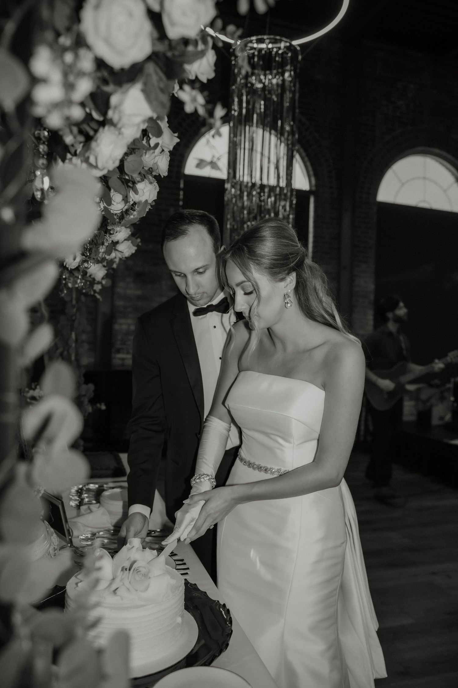Bride and groom cutting wedding cake together in black and white photo with floral arrangements.