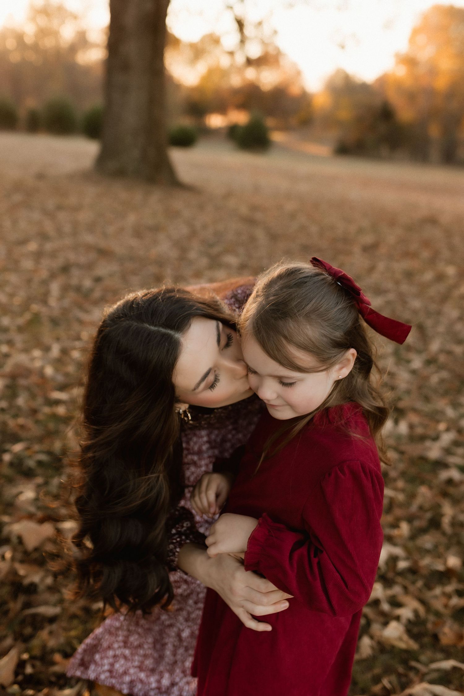 Mother tenderly kisses her young daughter in a red dress during a golden hour autumn outdoor portrait session.