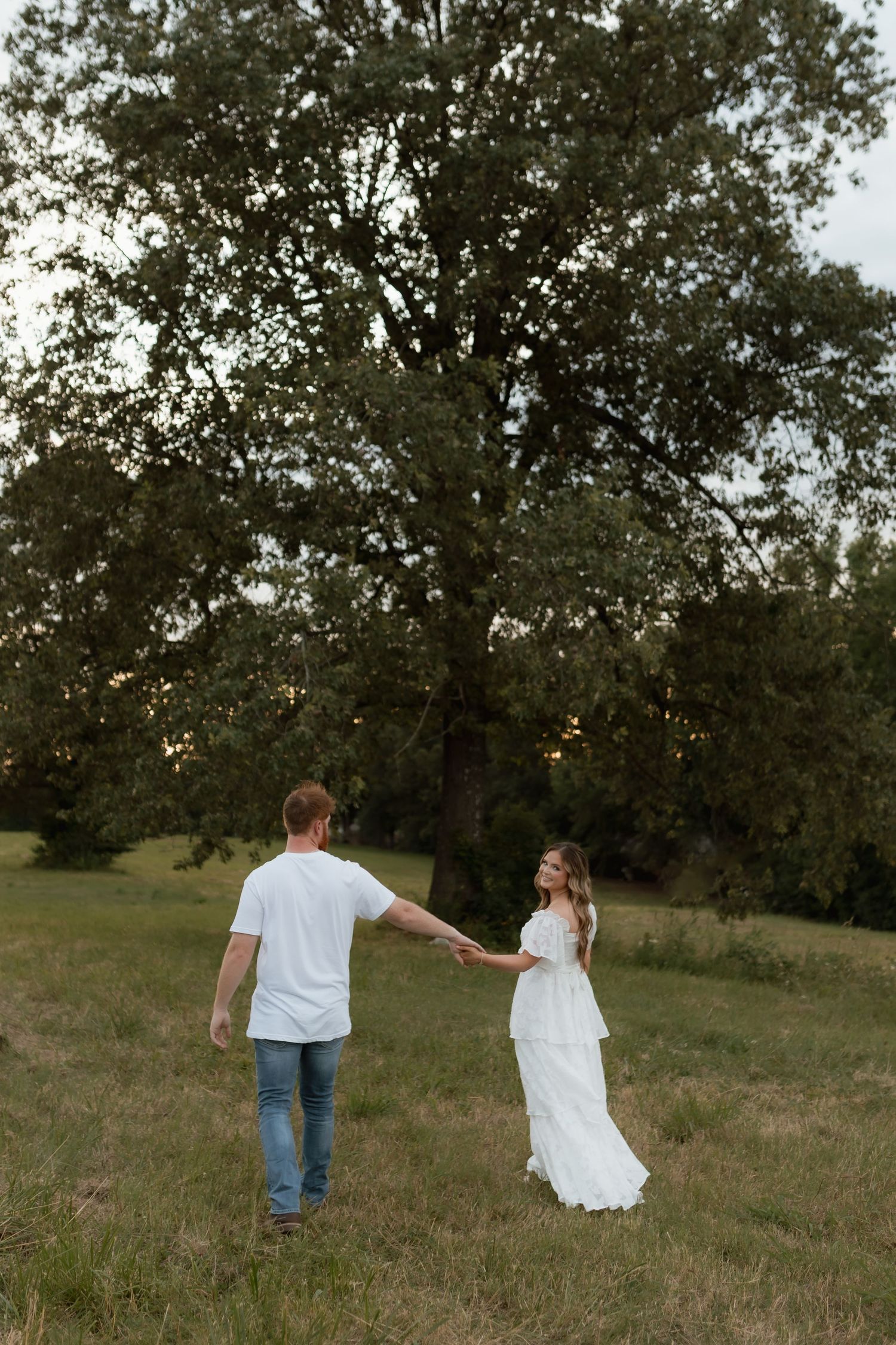 A couple holds hands walking toward a large tree in a grassy field at dusk.