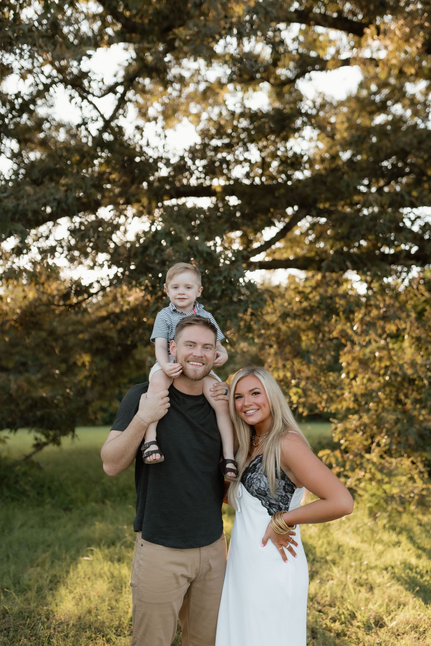Happy family with toddler on fathers shoulders posing under large tree in golden hour light.