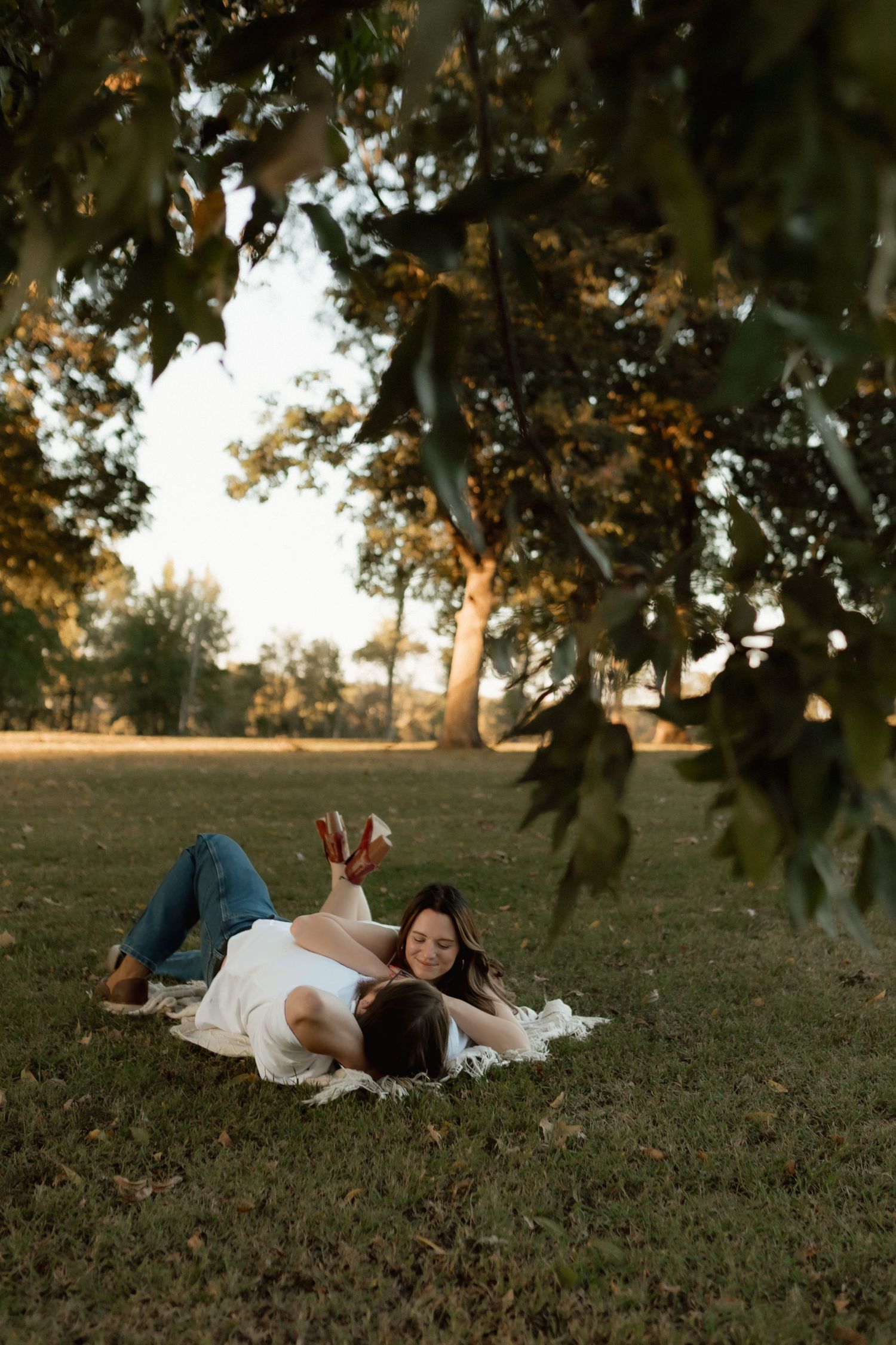 A couple relaxes on a blanket under a tree in a park at golden hour, laughing and enjoying each others company.