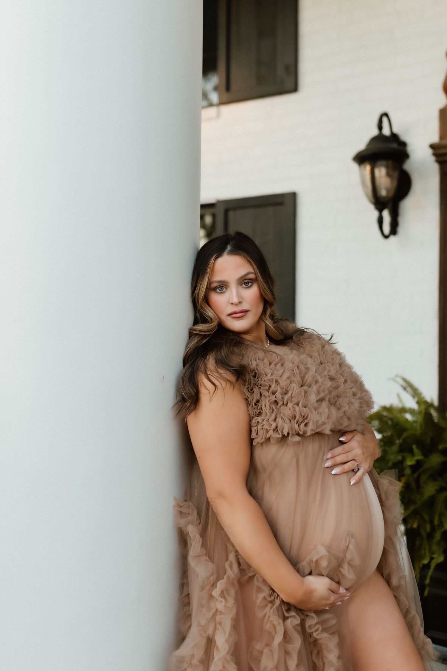 Pregnant woman in elegant tan tulle and fur maternity gown leaning against white column at golden hour.