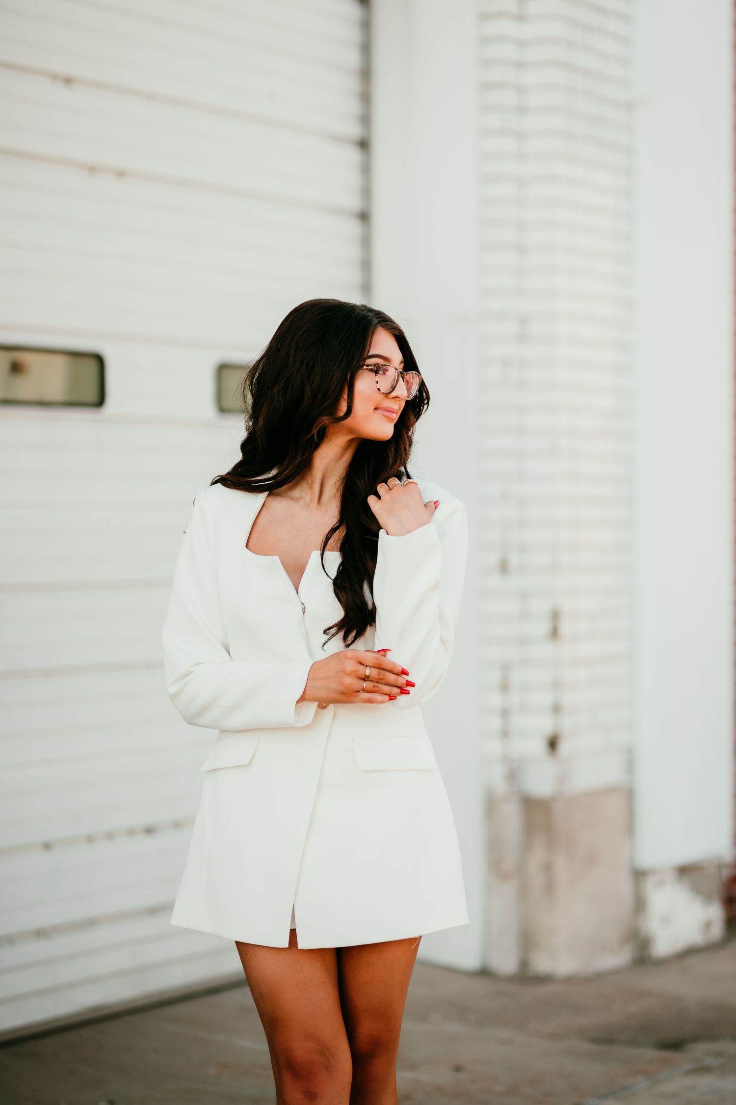 Brunette woman in white blazer dress posing outside white building.
