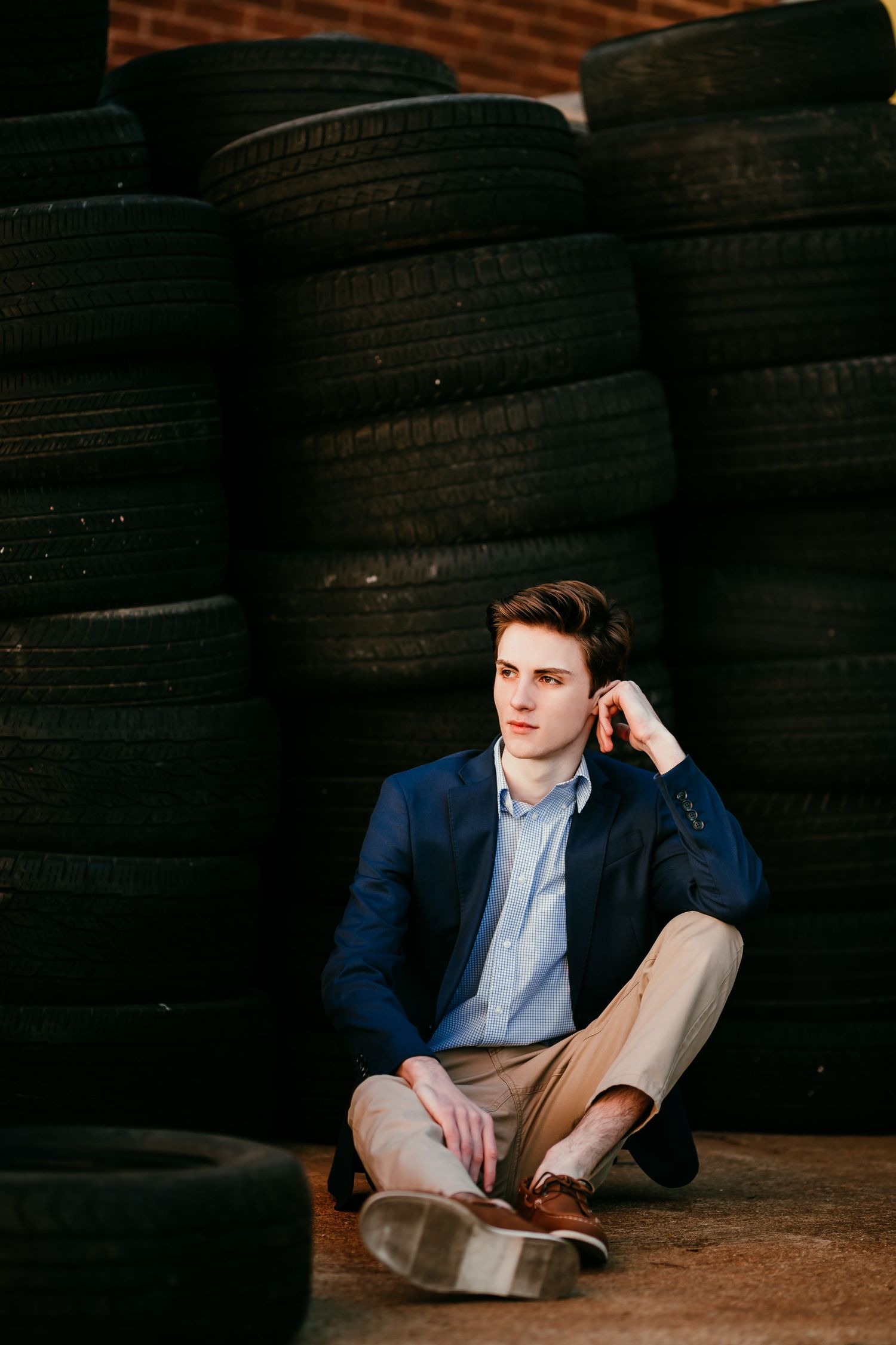 Young man in blue blazer sitting on ground against stacked tires in urban setting.