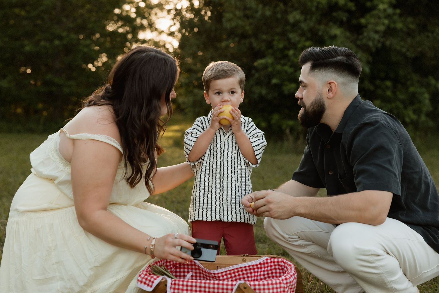 Young family with toddler eating fruit beside red gingham picnic basket in park at sunset.