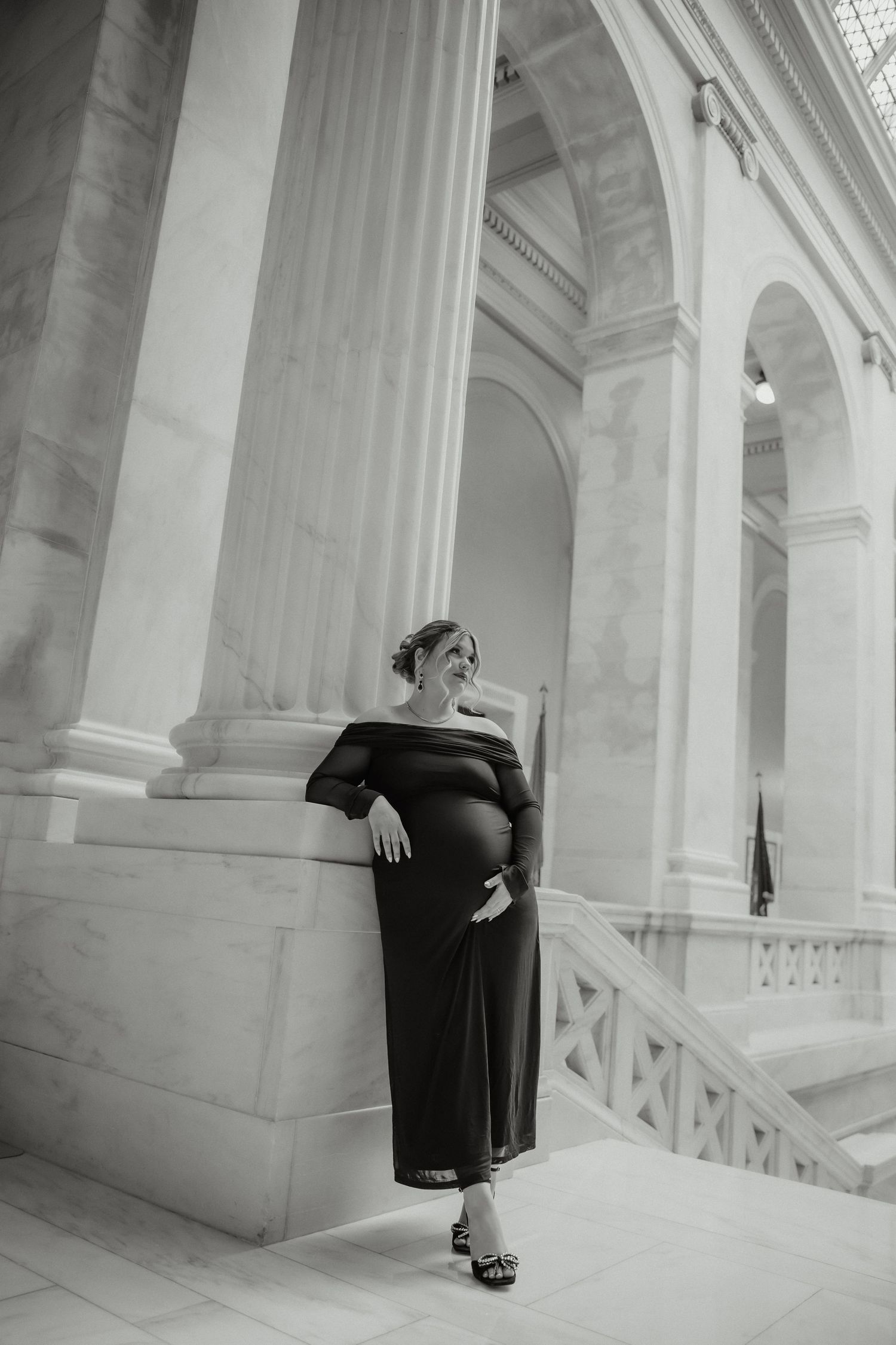 Pregnant woman in black off-shoulder dress leaning against marble column in grand interior, monochrome.