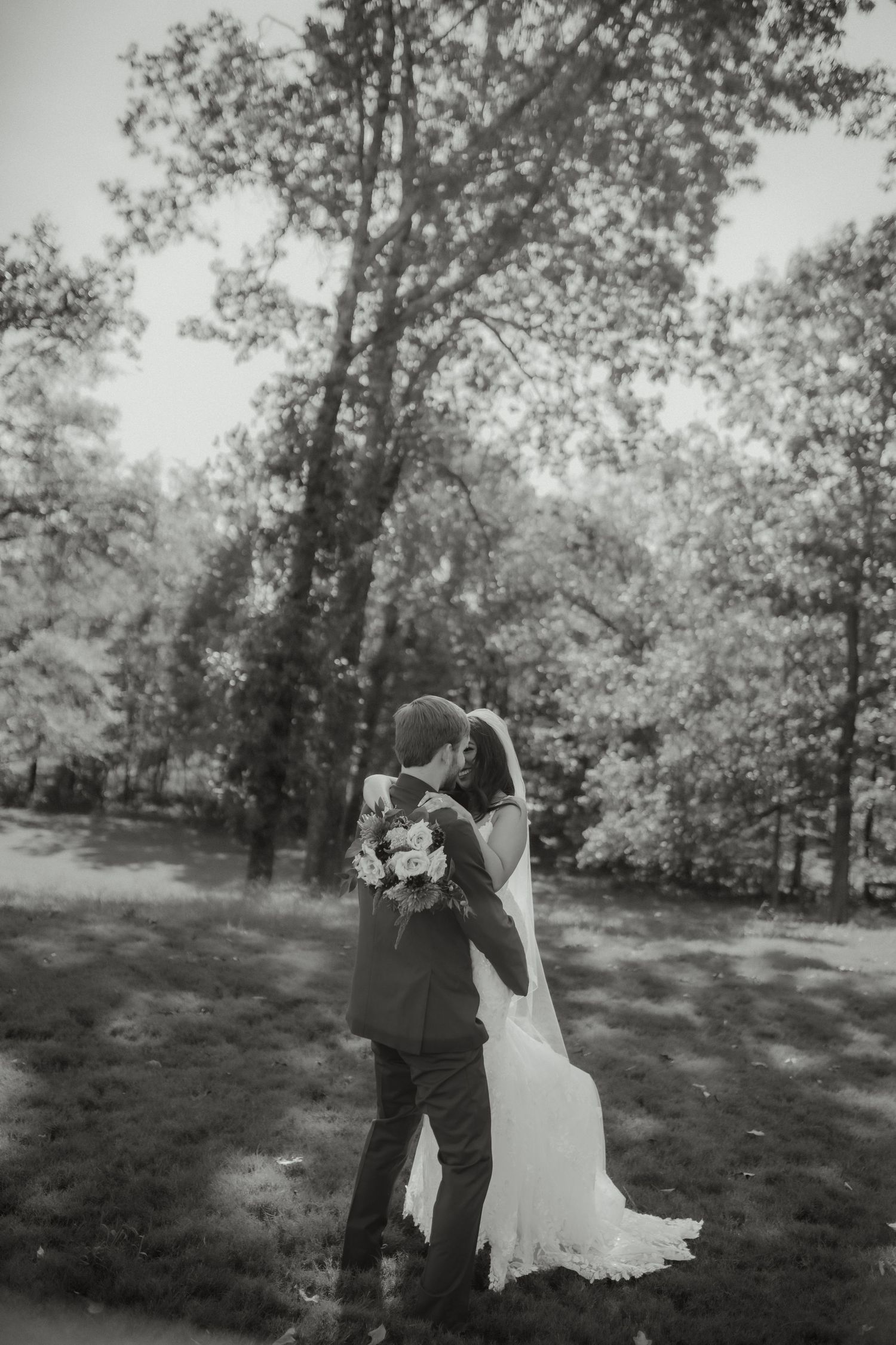 Black and white romantic photo of bride and groom embracing in a lush forest setting.