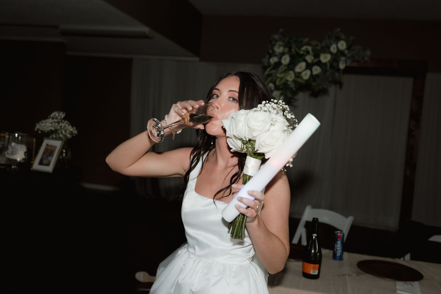 Bride playfully poses with her white bouquet and a bottle at the wedding reception table.