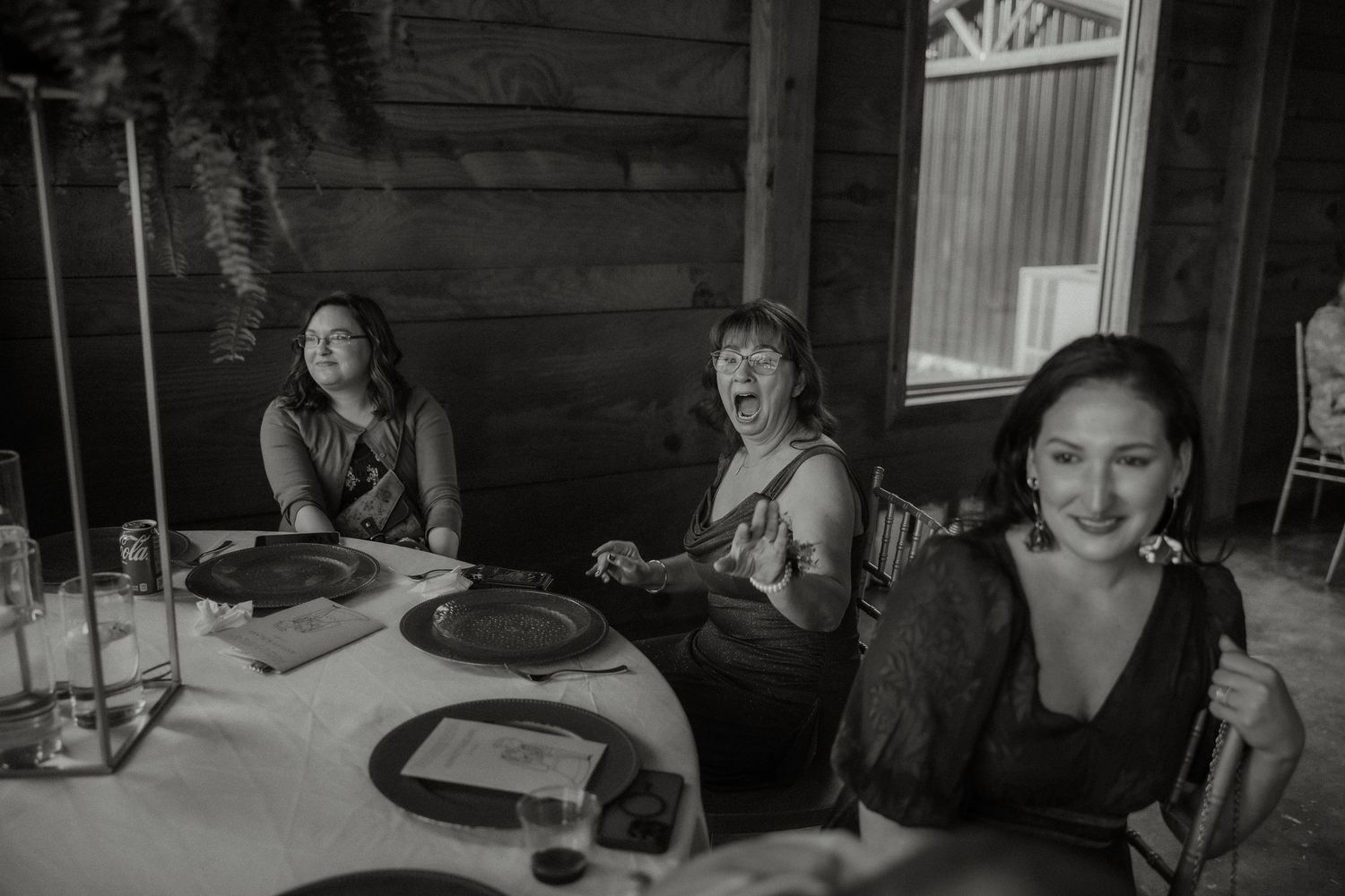 Black and white photo of three women laughing at wedding reception dinner table, candid moment.