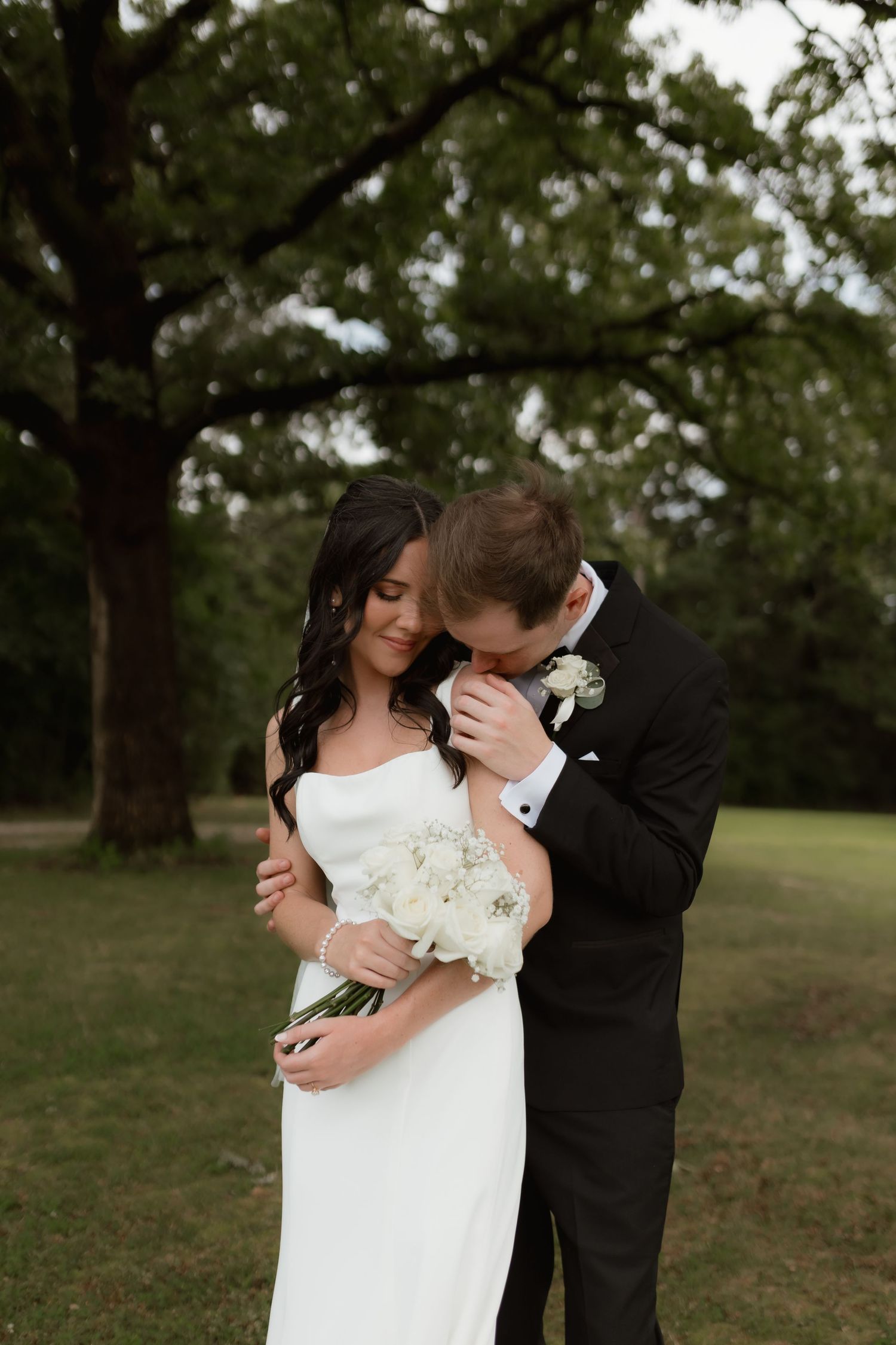 Groom tenderly embraces bride holding a white bouquet outdoors beneath towering oak trees.