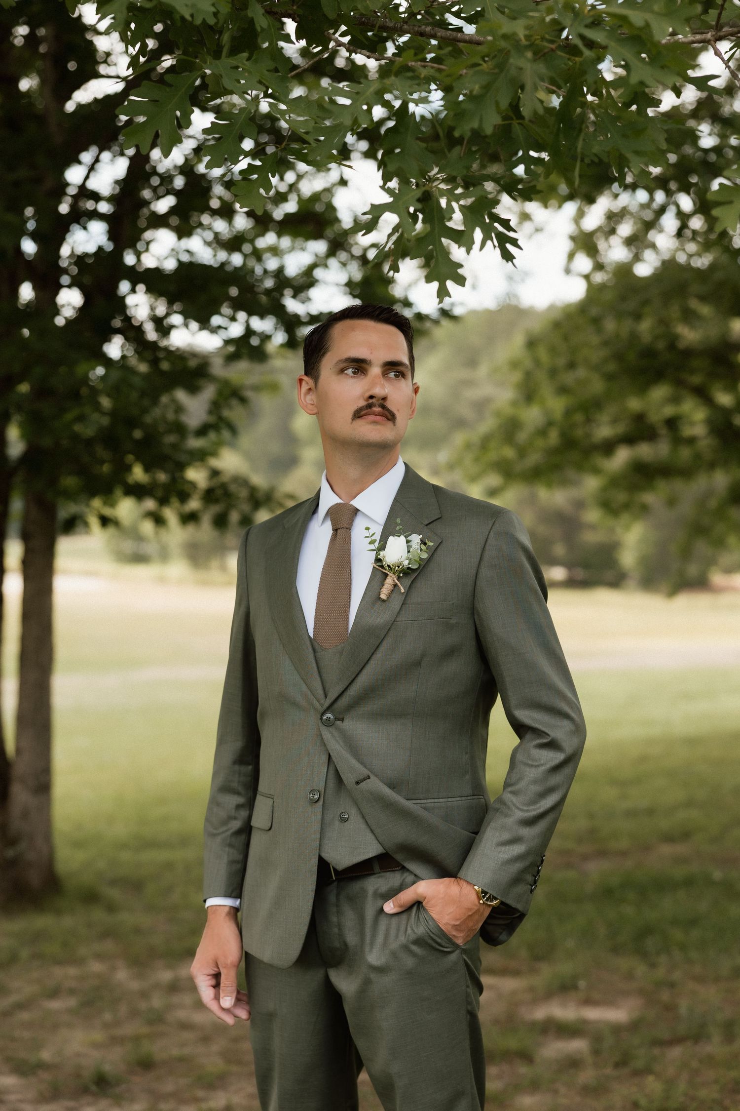Handsome groom in olive green three-piece suit with boutonniere standing under tree outdoors.