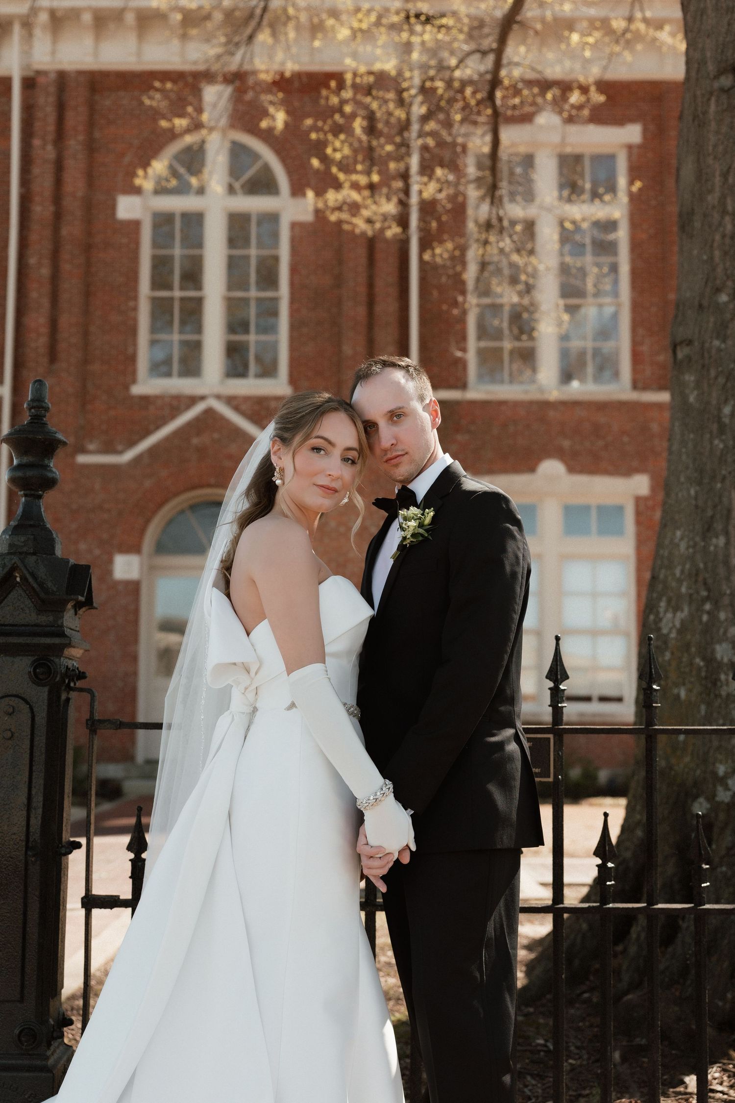 Elegant bride and groom pose hand in hand before a historic brick building in golden light.
