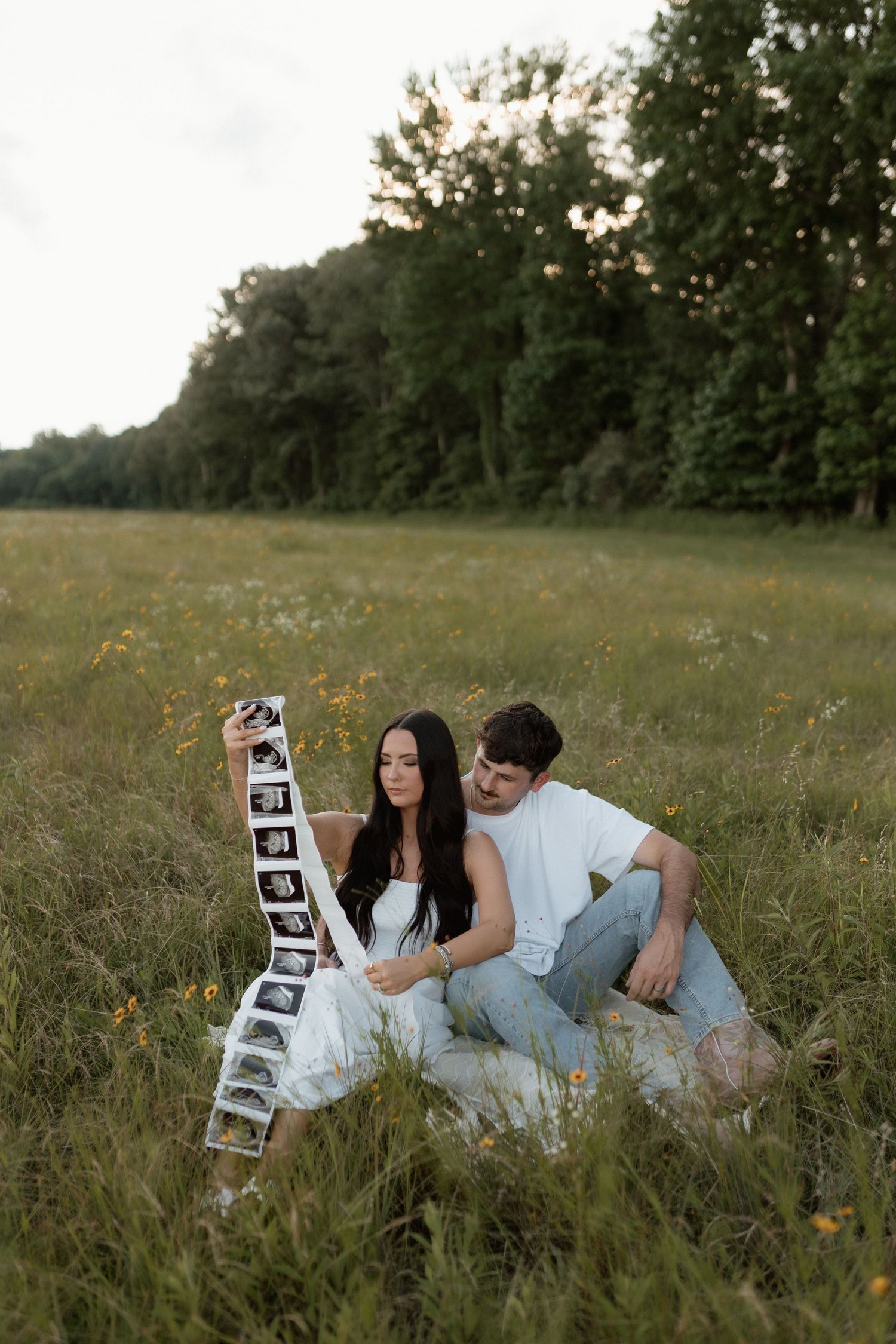 Pregnant couple sitting in a field examining a strip of ultrasound images in the golden hour light surrounded by wildflowers.