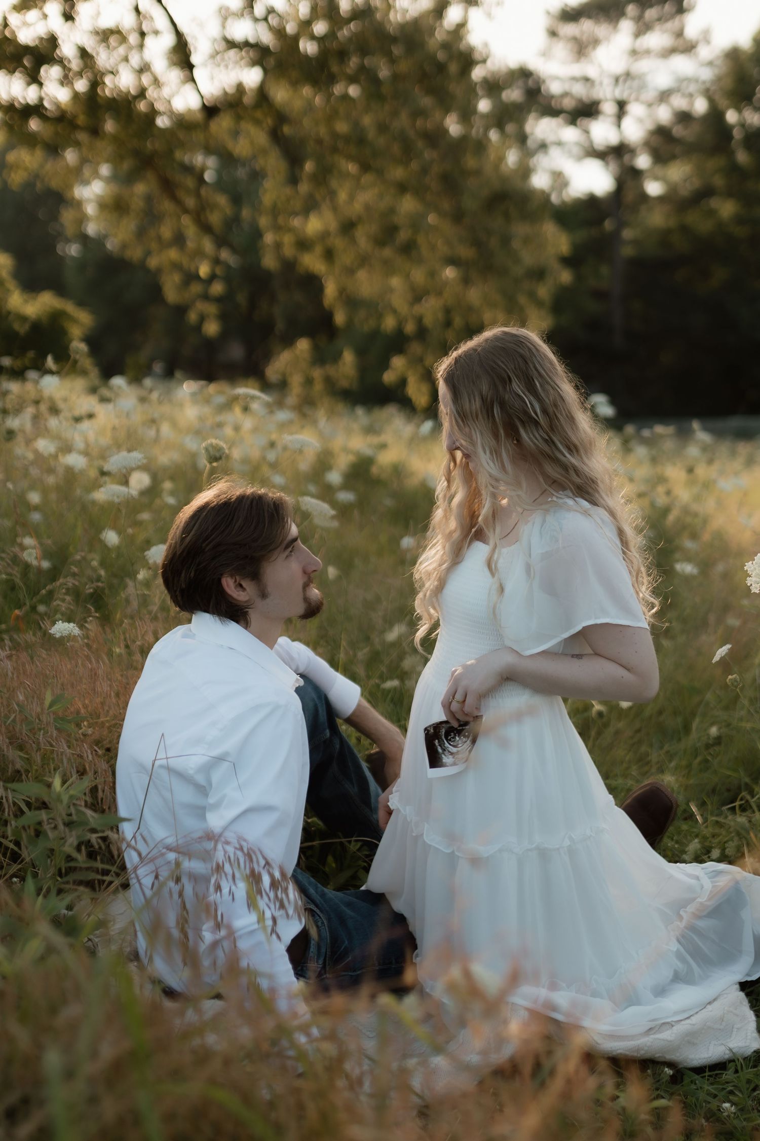 Man sitting in a wildflower meadow looking up at pregnant woman in white dress holding ultrasound photo at golden hour.