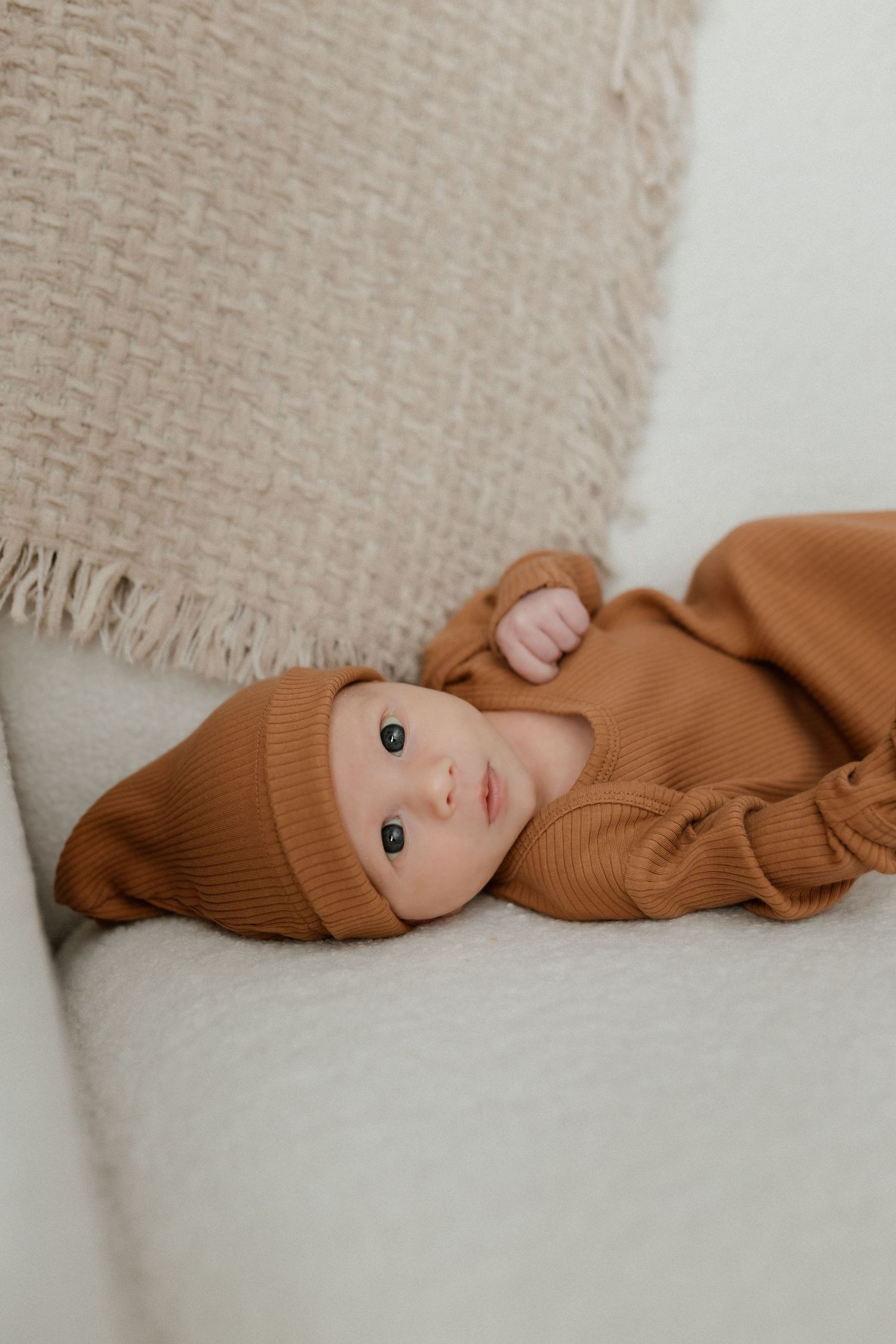 Alert newborn baby in a rust-colored knit outfit and hat lies on a cream sofa with a textured blanket.