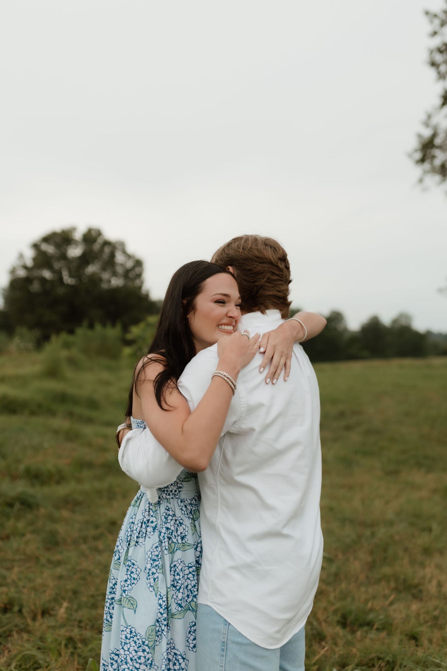 A joyful woman in a floral dress hugs a man in a white shirt tightly in an outdoor field setting.