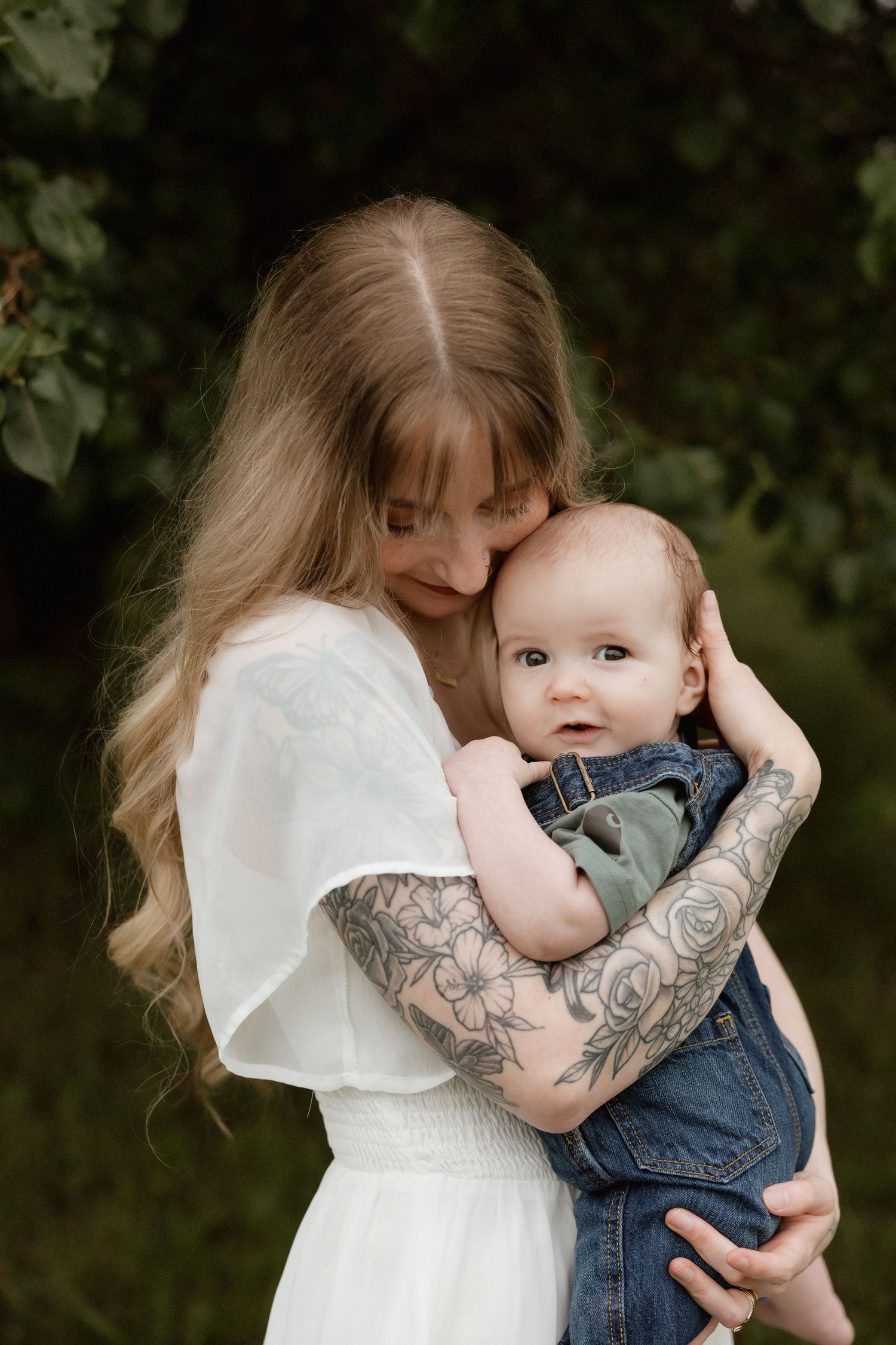 Blonde mother in white dress tenderly embracing infant in denim outfit with tattooed arms.