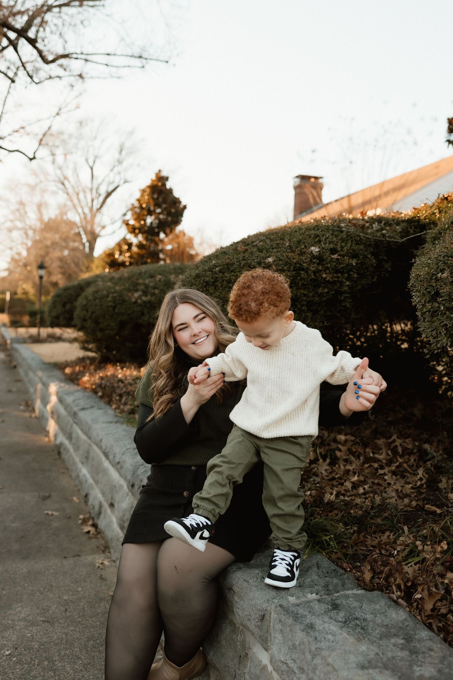 Young mother in black dress holding toddler boy's hands on stone wall in autumn park.