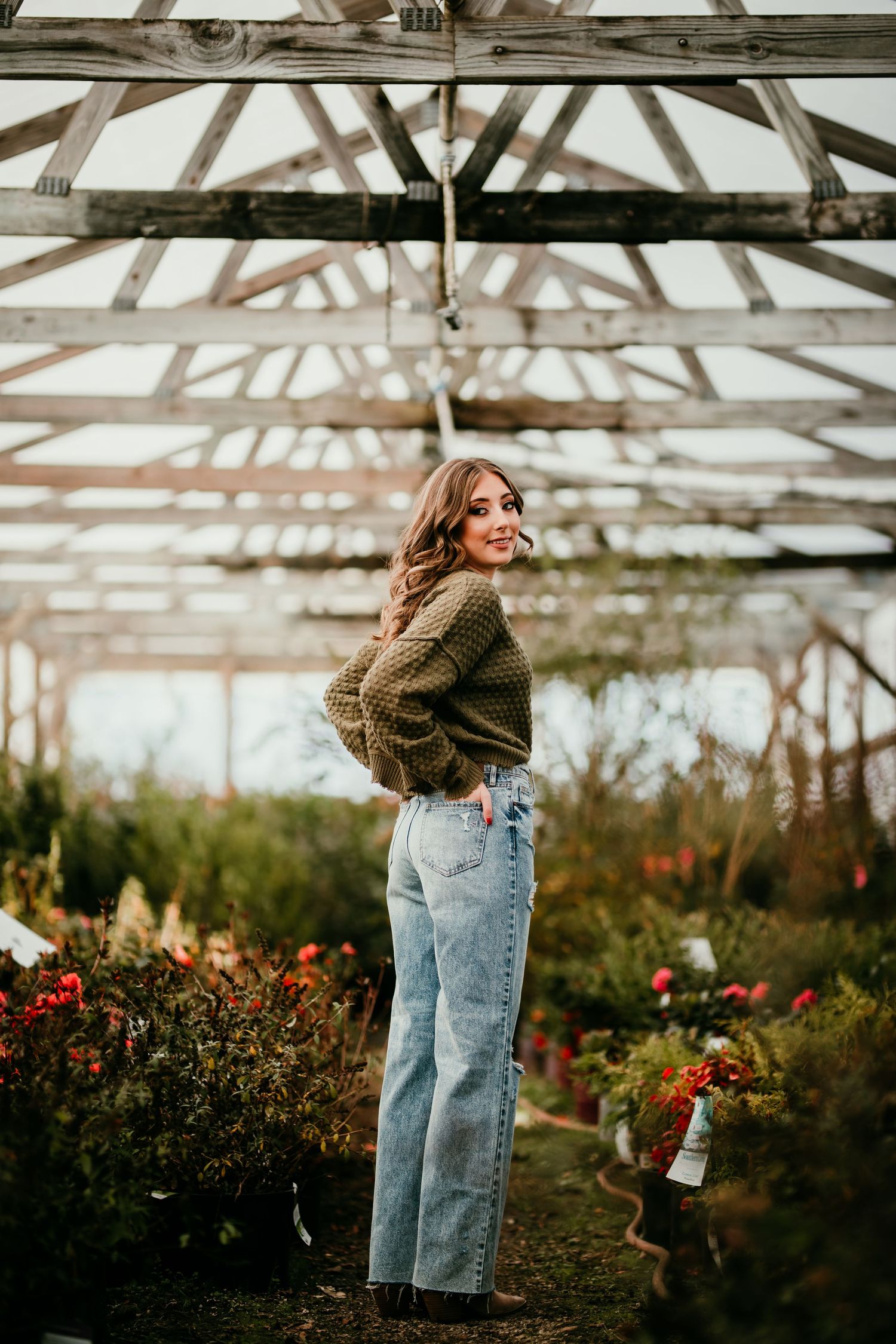 Young woman in olive sweater and wide-leg jeans poses in rustic greenhouse surrounded by plants.