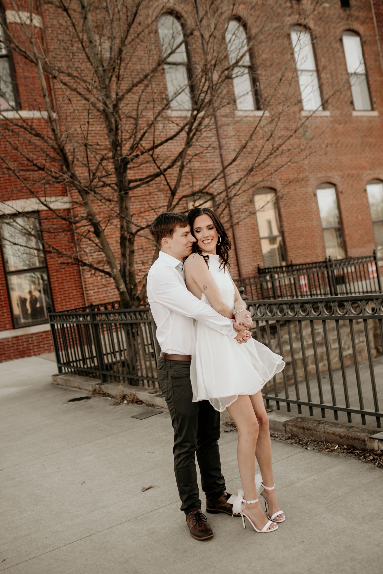 Couple embracing and smiling in front of historic brick building during urban winter engagement session.