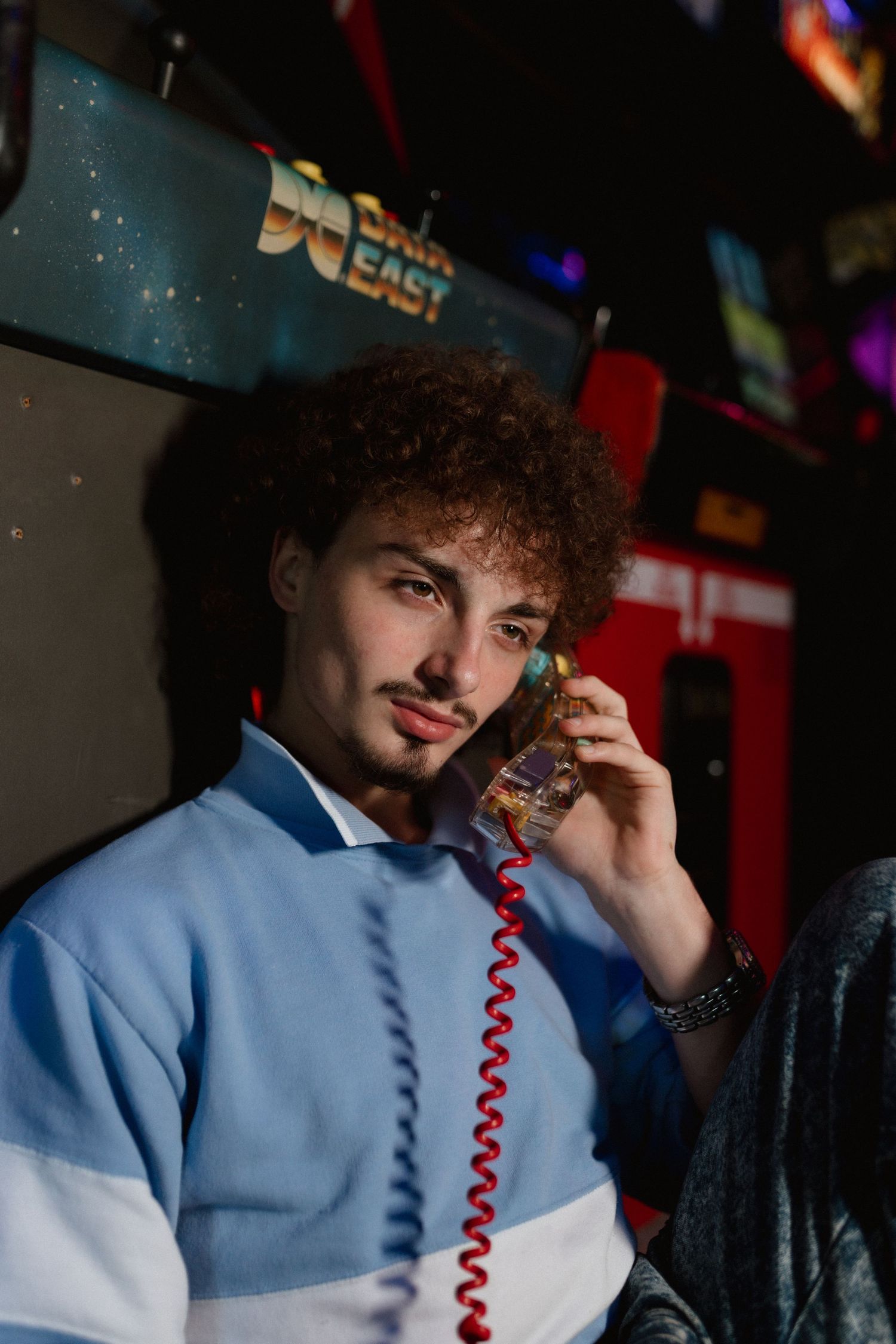 Young man with curly hair holding vintage phone in retro arcade, blue sweatshirt.