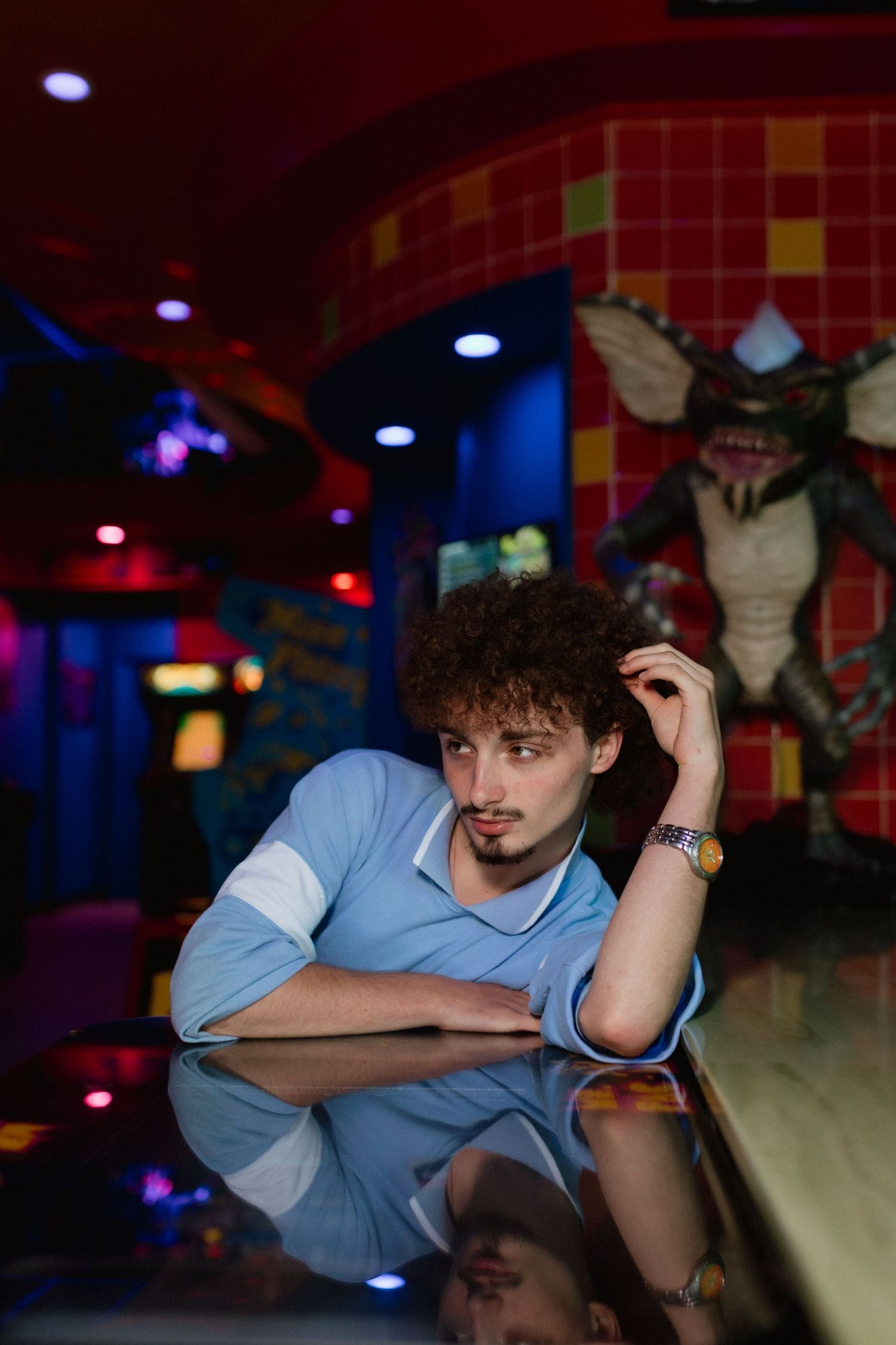 Young man with curly hair resting on reflective surface in colorful retro arcade bar.