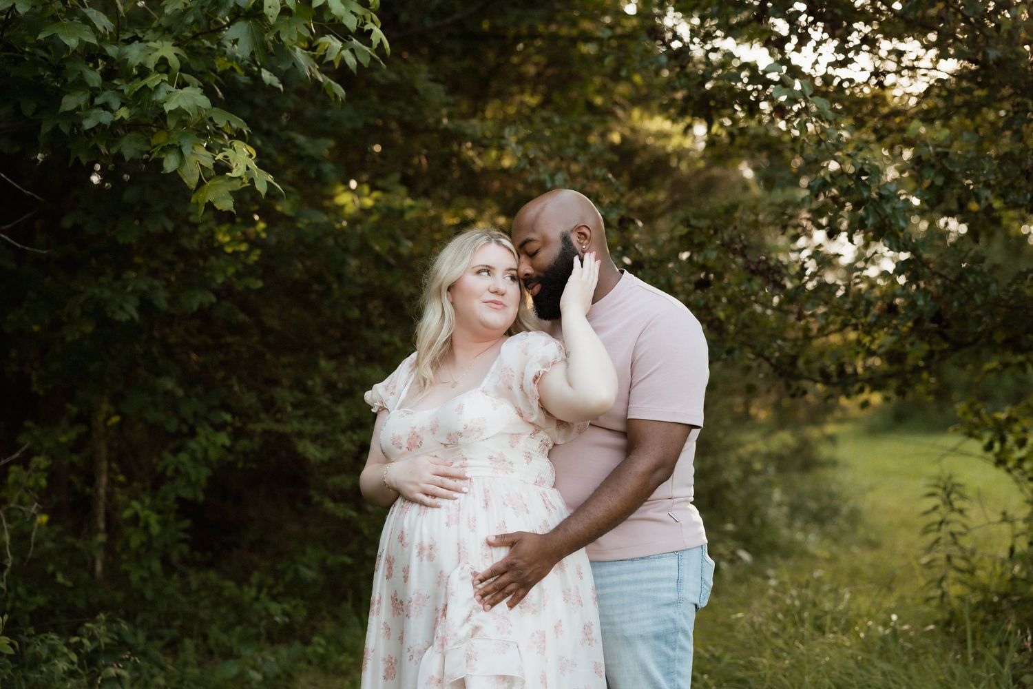 Pregnant couple embracing in a sunlit garden, woman in floral maternity dress holding her belly.