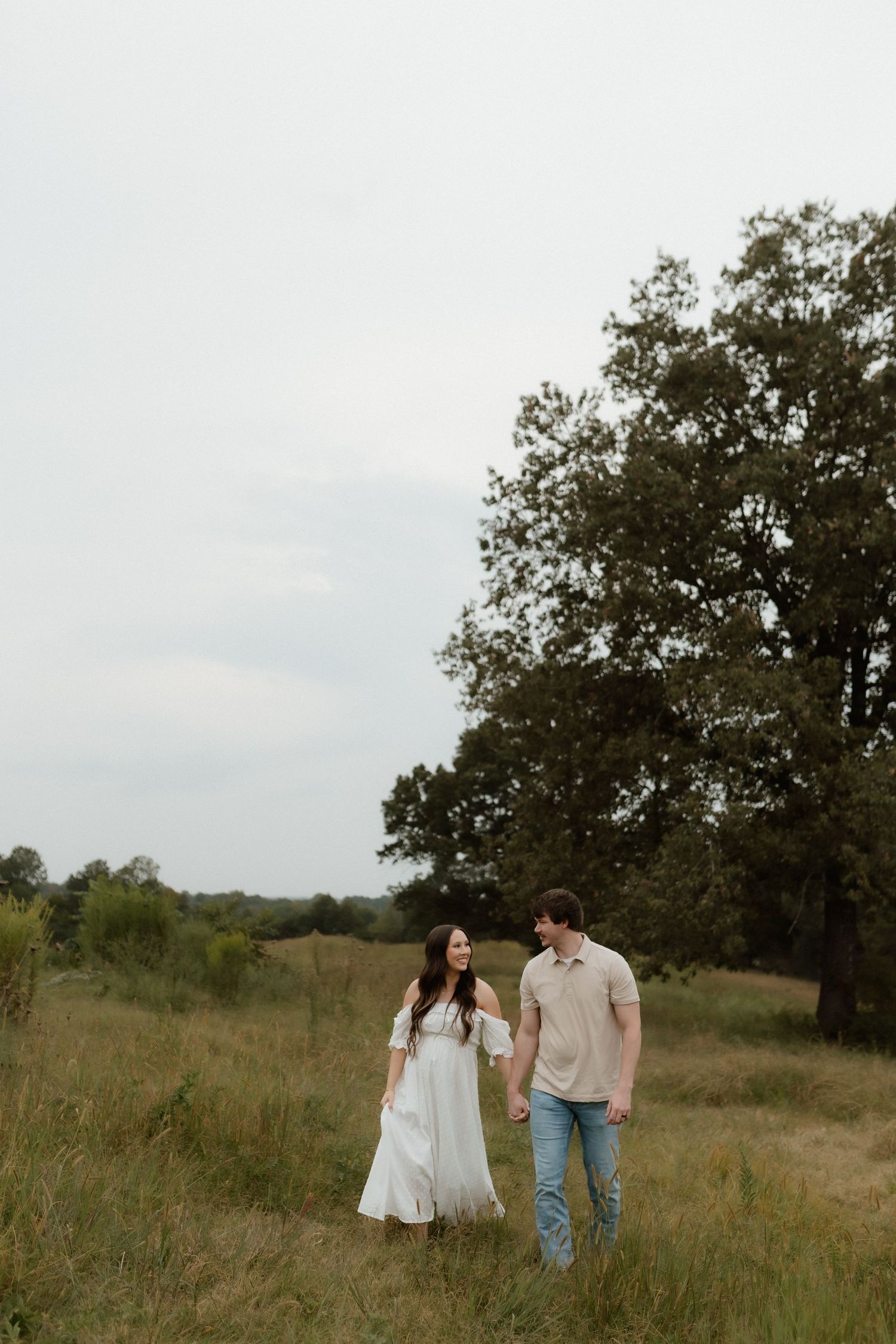 Expecting couple walks hand-in-hand through open grassy field with tall trees behind them.