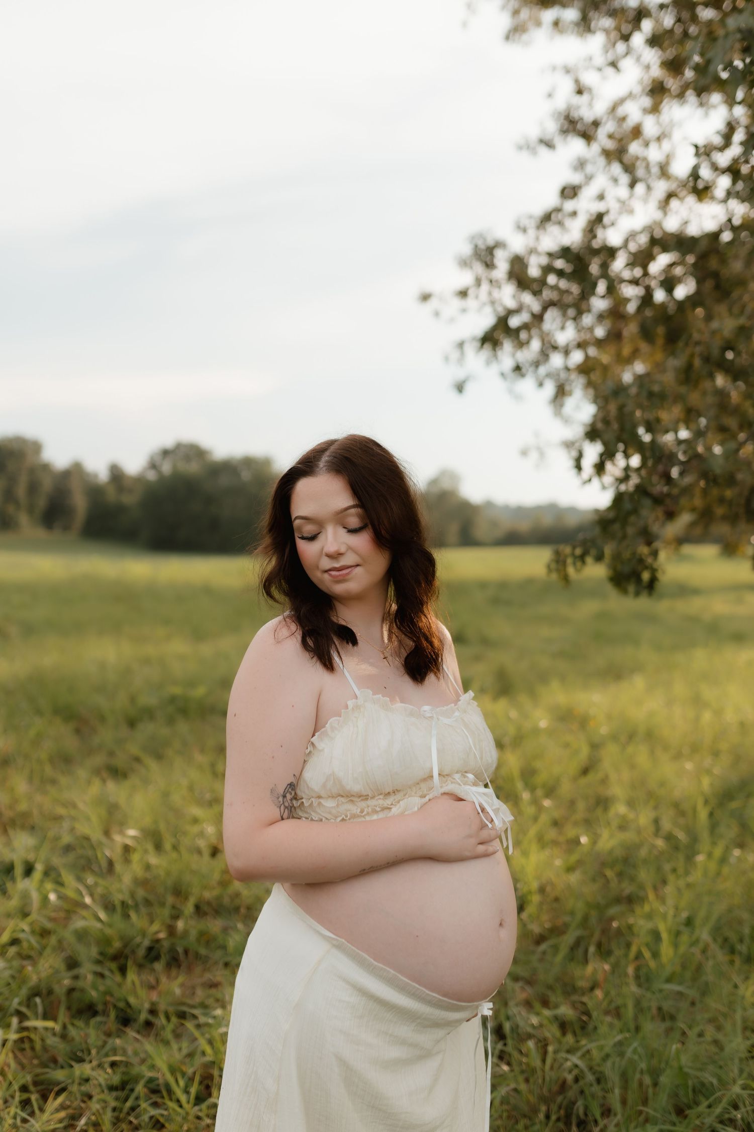 Pregnant woman in cream dress standing in green meadow, looking down at bare baby bump.