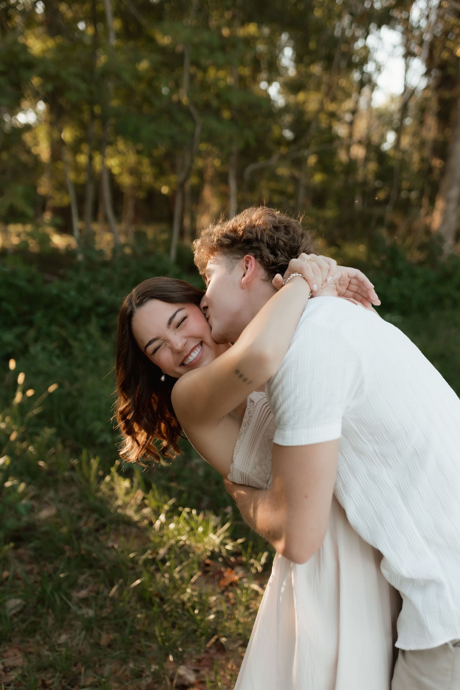Man kissing laughing woman on cheek during outdoor couples photo session in green forest setting.
