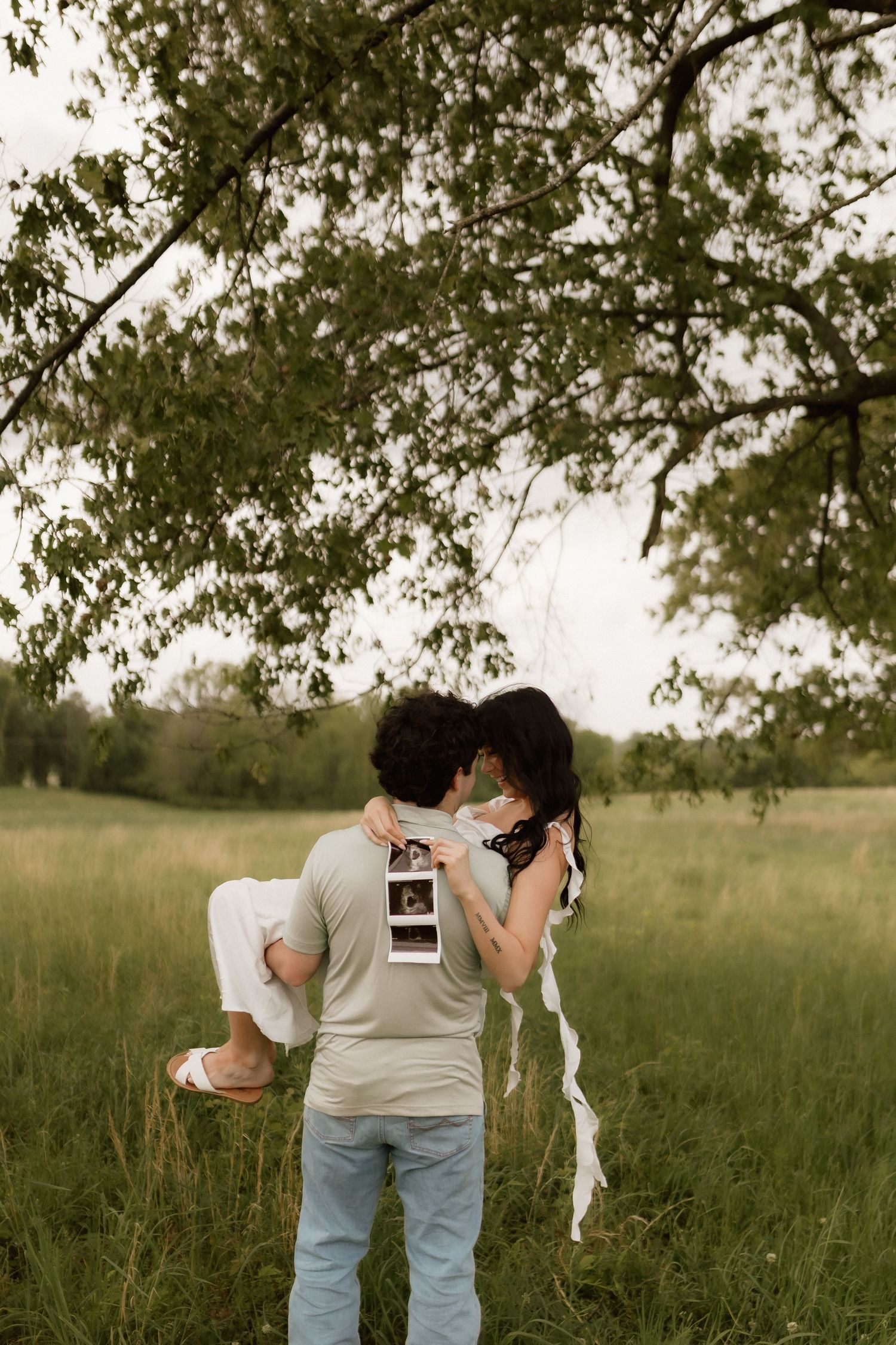 Man lifting pregnant woman in white dress while kissing, holding ultrasound photos in lush green field.