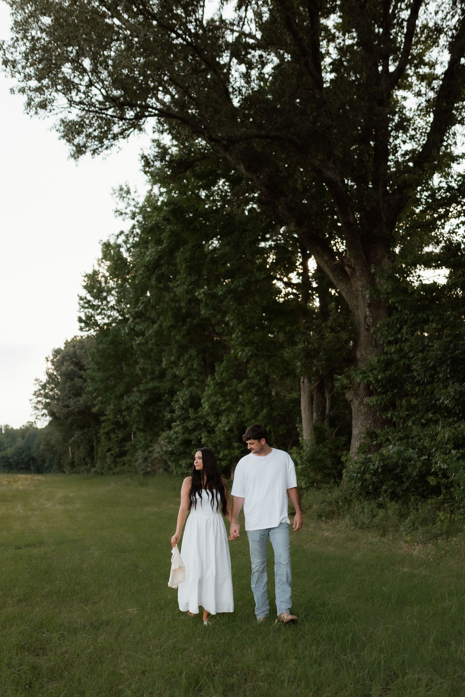 Couple walking hand in hand through a lush green field at dusk, woman in white dress, man in white shirt and jeans.