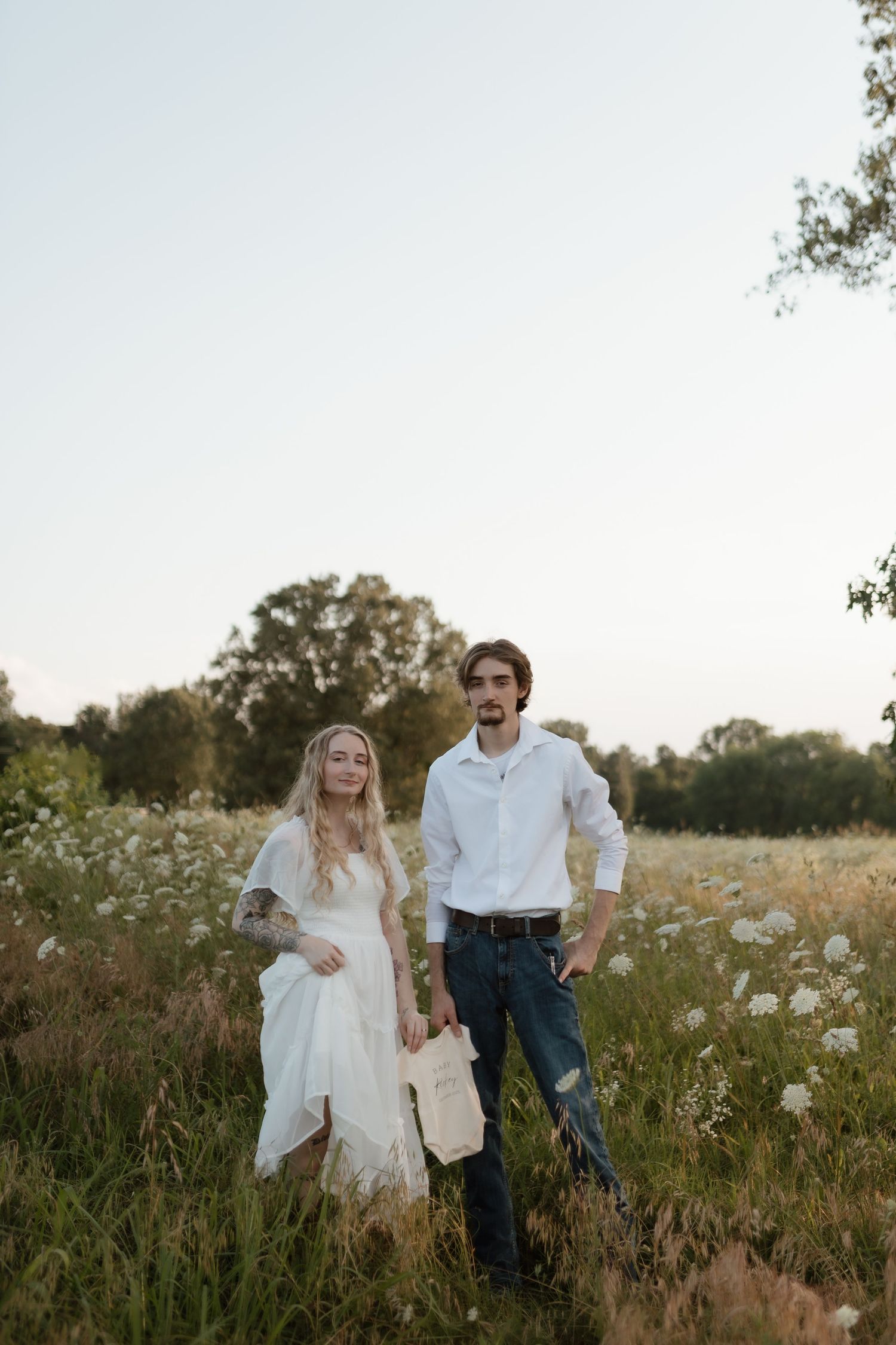 Tattooed blonde woman in white dress and bearded man in white shirt walking through a wildflower meadow at sunset.