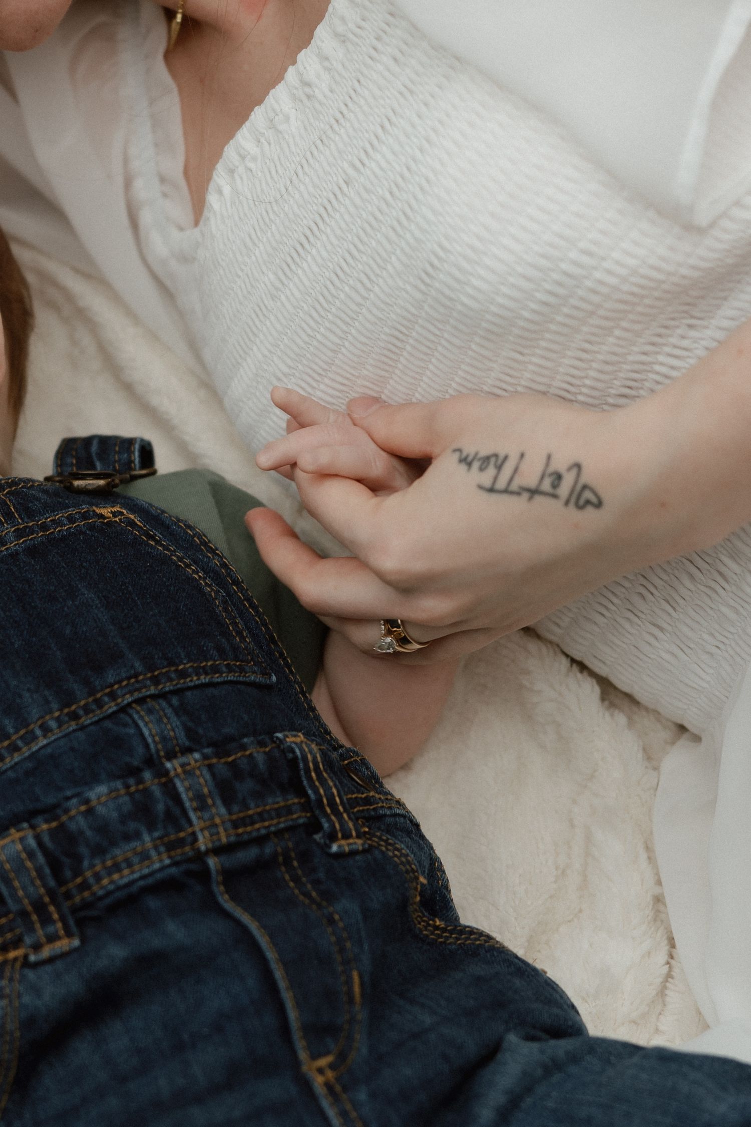 Close-up of tattooed mother holding baby's hands in denim overalls.