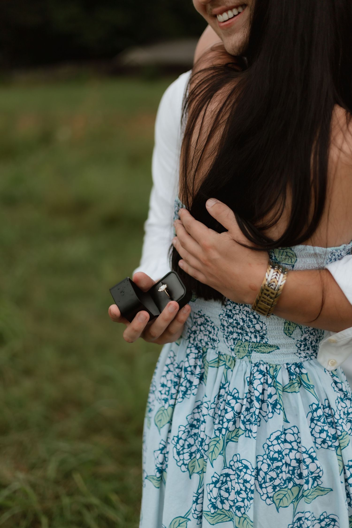 A close-up of a couple embracing outdoors, the man holding a ring box behind the smiling woman.