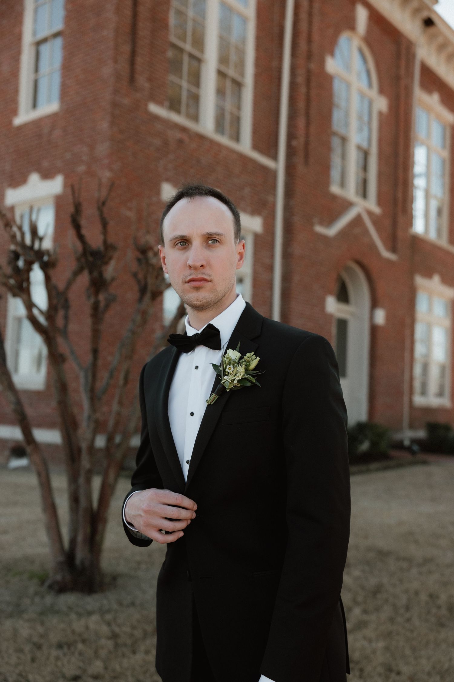 Dapper groom in black tuxedo with boutonniere stands before a brick building.