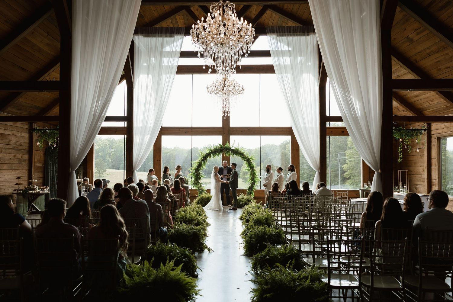 Wedding ceremony inside rustic barn venue with chandelier, greenery arch, and guests seated in rows.