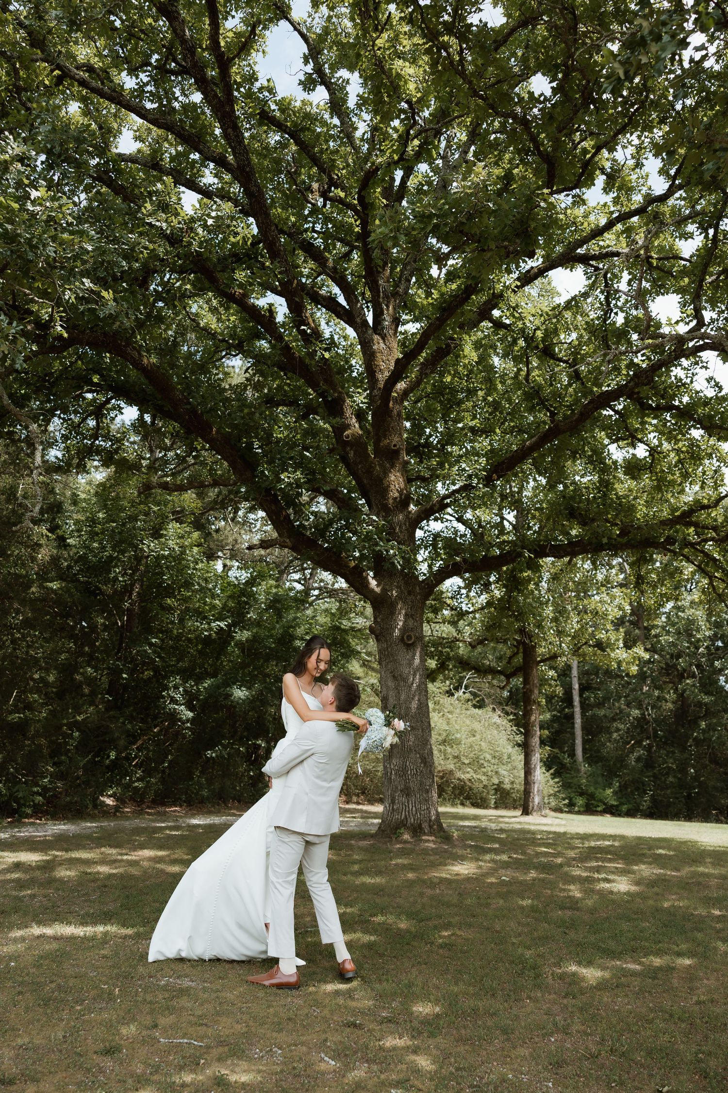 Groom lifting bride in white dress outdoors under large oak tree on wedding day.