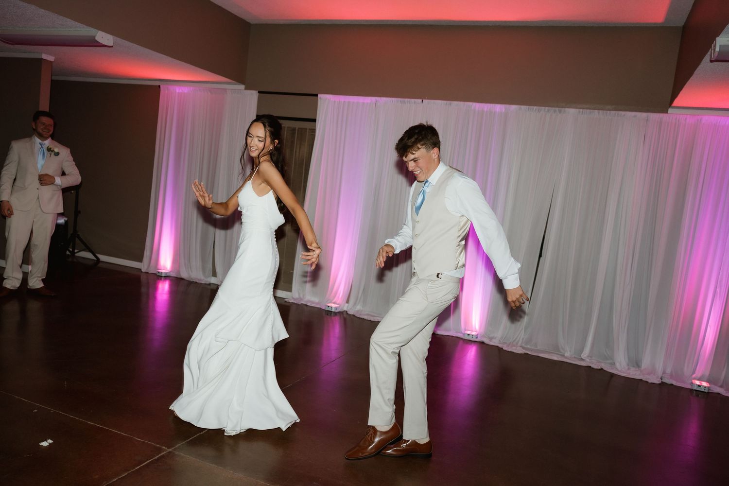 Bride in fitted gown and groom in white suit dancing joyfully at wedding reception.