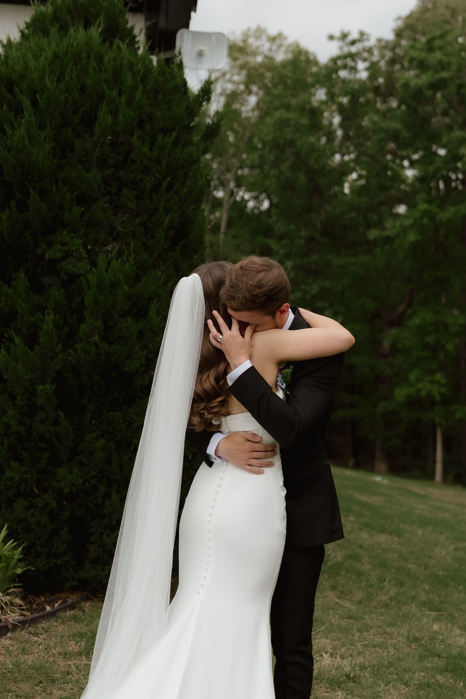 Bride and groom sharing first look embrace outdoors, bride in fitted gown with long veil.