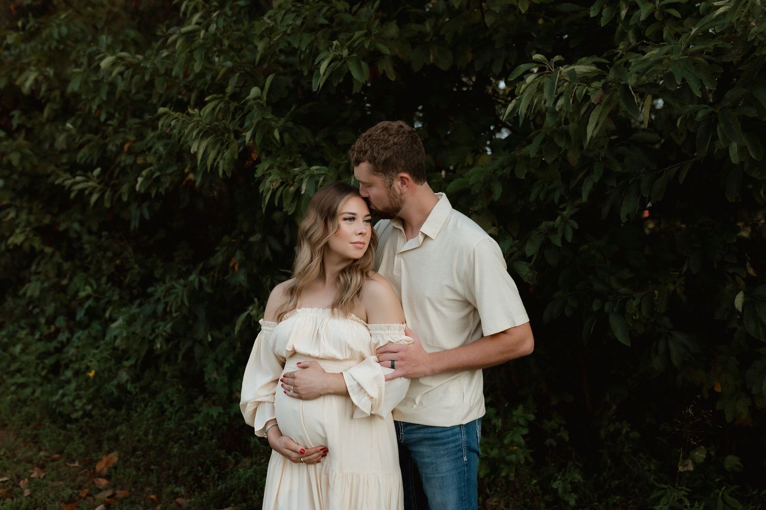 Expecting couple standing together in front of lush greenery, man tenderly kissing pregnant woman's head outdoors.