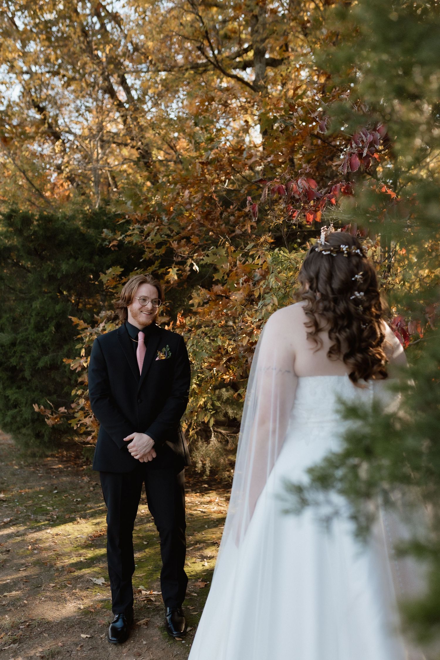 Groom in black tuxedo smiling at bride in white wedding dress during first look in autumn garden setting.