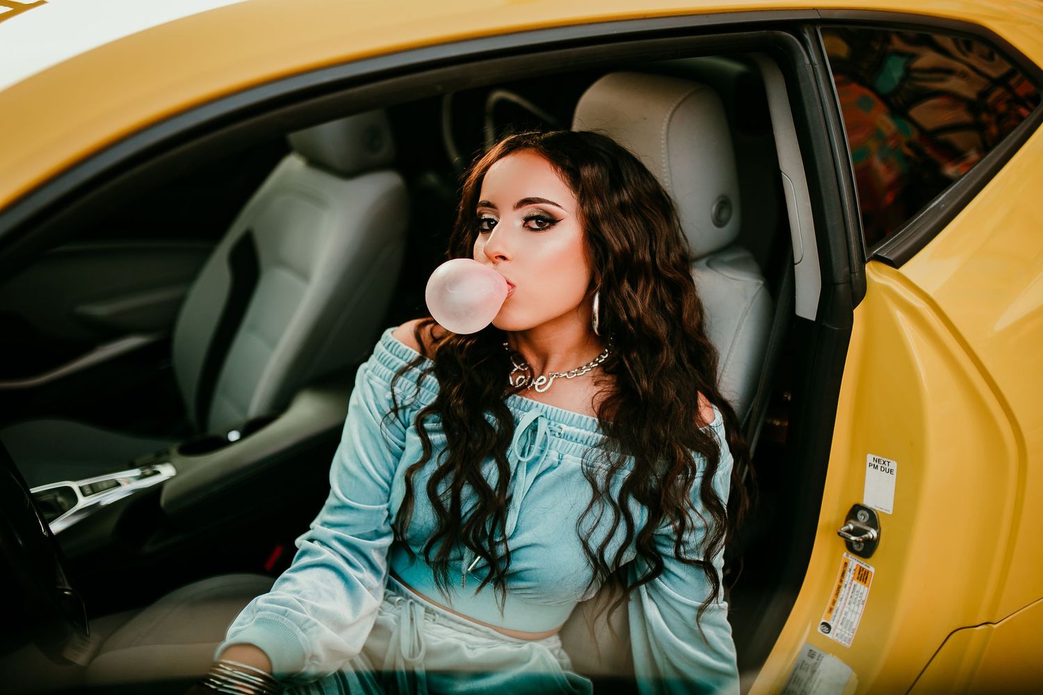 Woman in blue velvet outfit blowing bubble gum sitting in yellow sports car.