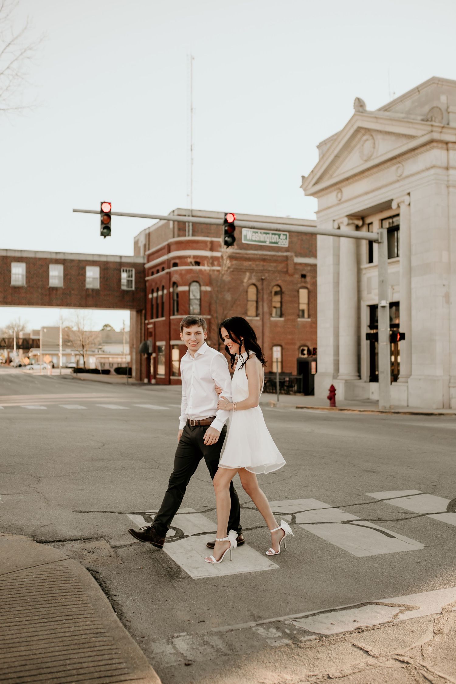 Engaged couple crossing downtown street intersection, woman in white mini dress and heels in golden hour light.