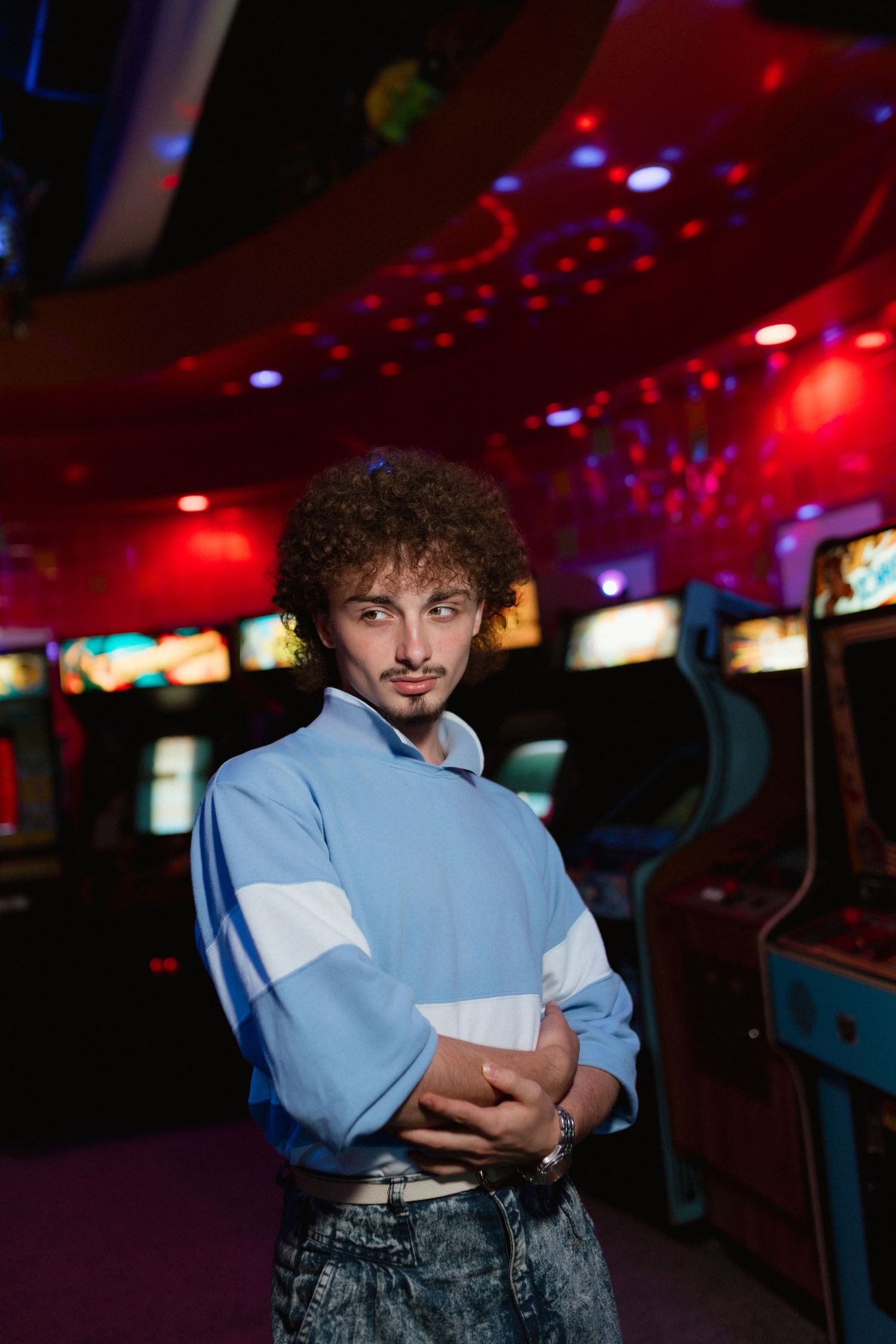 Curly-haired man in blue 80s-style sweatshirt posing in neon-lit retro arcade game room.